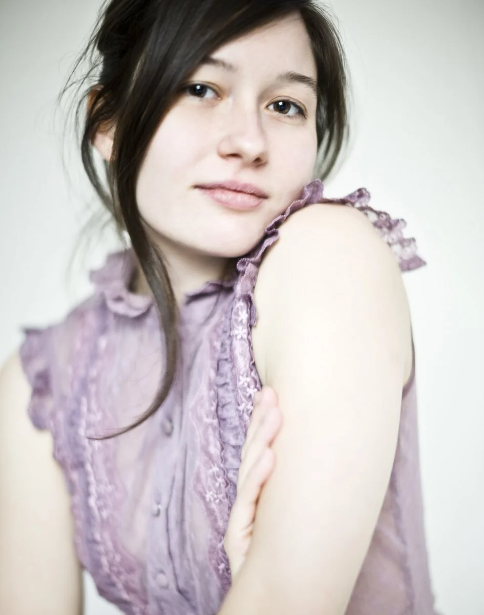 A young woman with dark hair and fair skin wearing a lilac sleeveless top, posing with her arm crossed over her shoulder in front of a plain background.