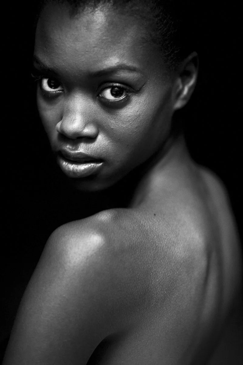 Close-up black and white portrait of a woman with smooth skin, looking directly at the camera, with her shoulder and upper arm visible.