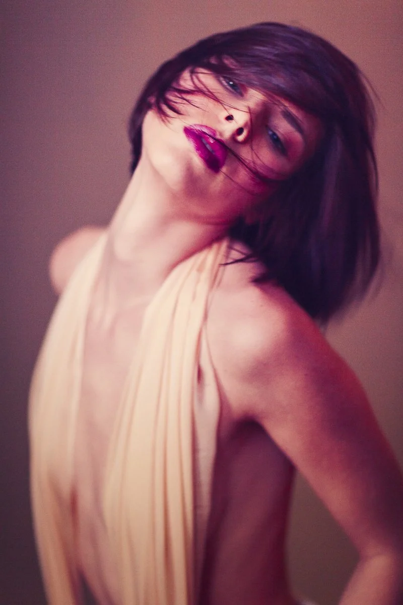 A woman with shoulder-length dark hair, wearing makeup with pink lipstick, poses with her head tilted back and eyes half closed, wearing a cream-colored sleeveless top.