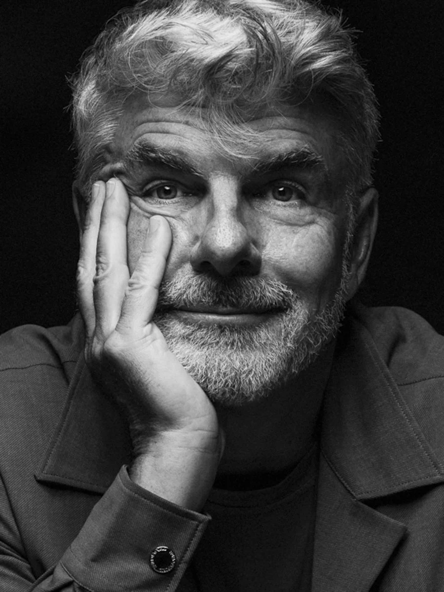 A black-and-white portrait of a middle-aged man with gray hair and a beard, resting his chin on his left hand, looking directly at the camera.