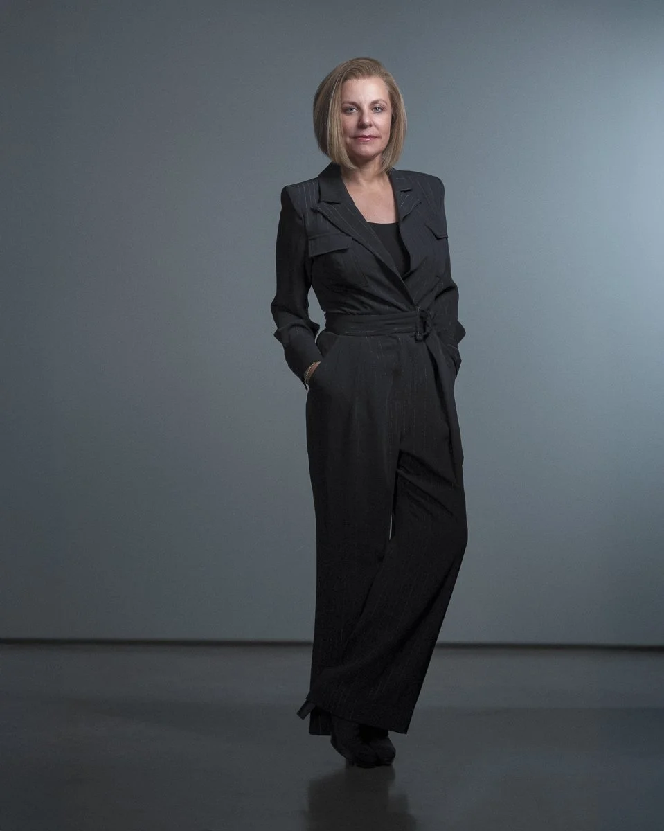 A woman in a black pinstripe suit standing against a plain gray background with hands in pockets.