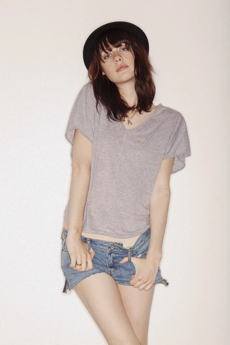 A young woman with dark hair, wearing a black hat, a gray t-shirt, and denim shorts, standing against a plain white background.