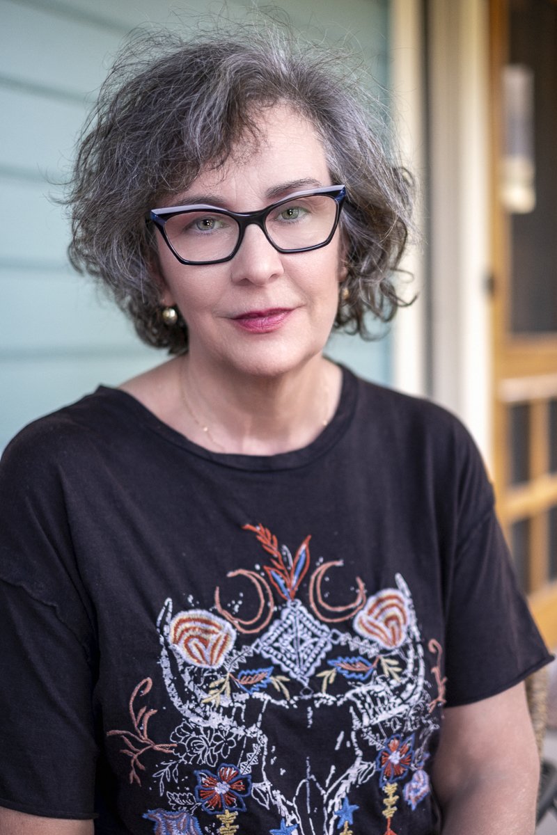 A woman with gray, curly hair and black-framed glasses, wearing a black embroidered T-shirt and earrings, sitting outdoors near a building with wooden elements.