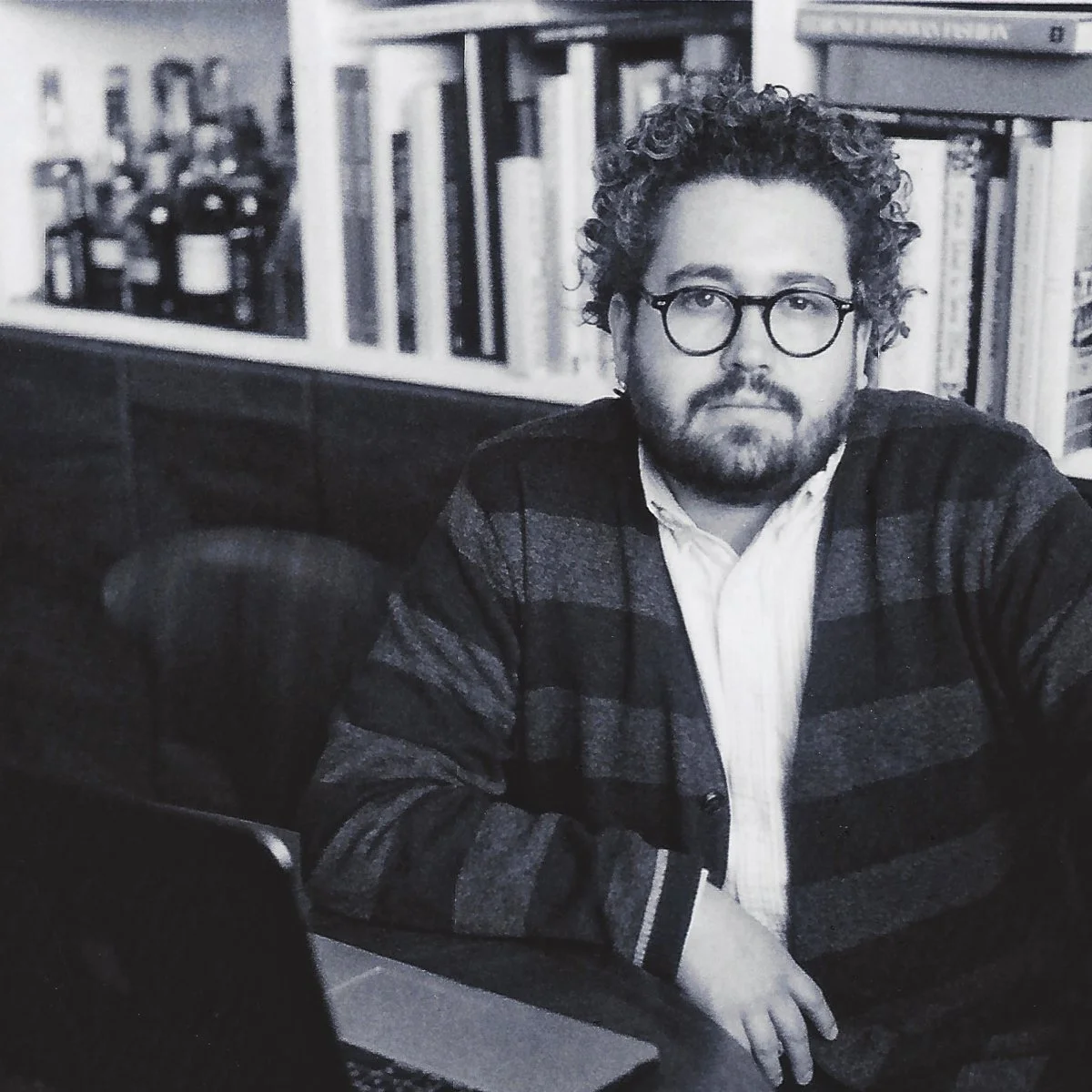 Black and white photo of a man with curly hair, glasses, and a beard, sitting at a table with bookshelves in the background.