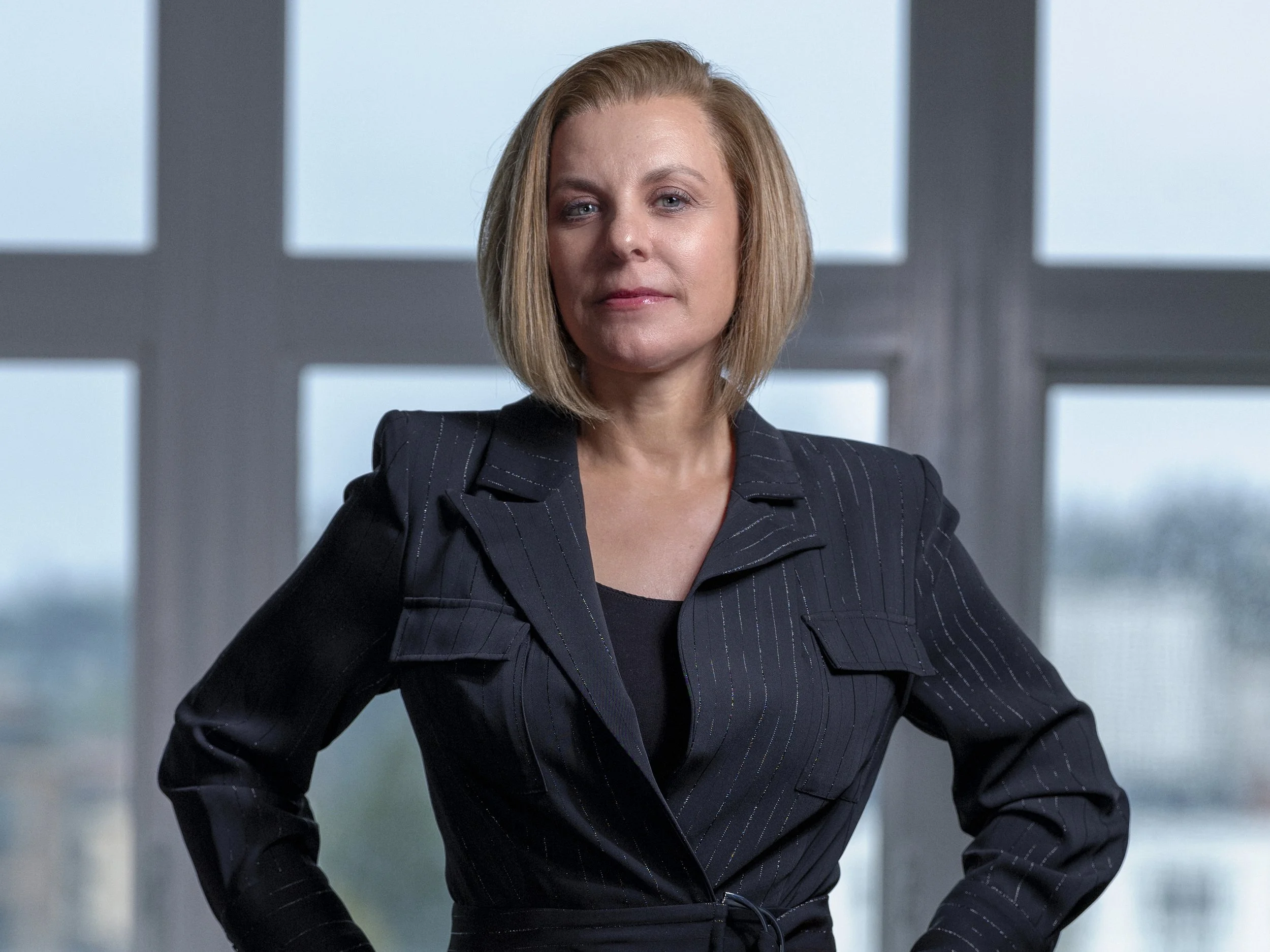 A woman with blonde hair styled in a bob, wearing a black pinstripe suit, standing confidently with her hand on her hip in an office or professional setting with a blurred geometric background.