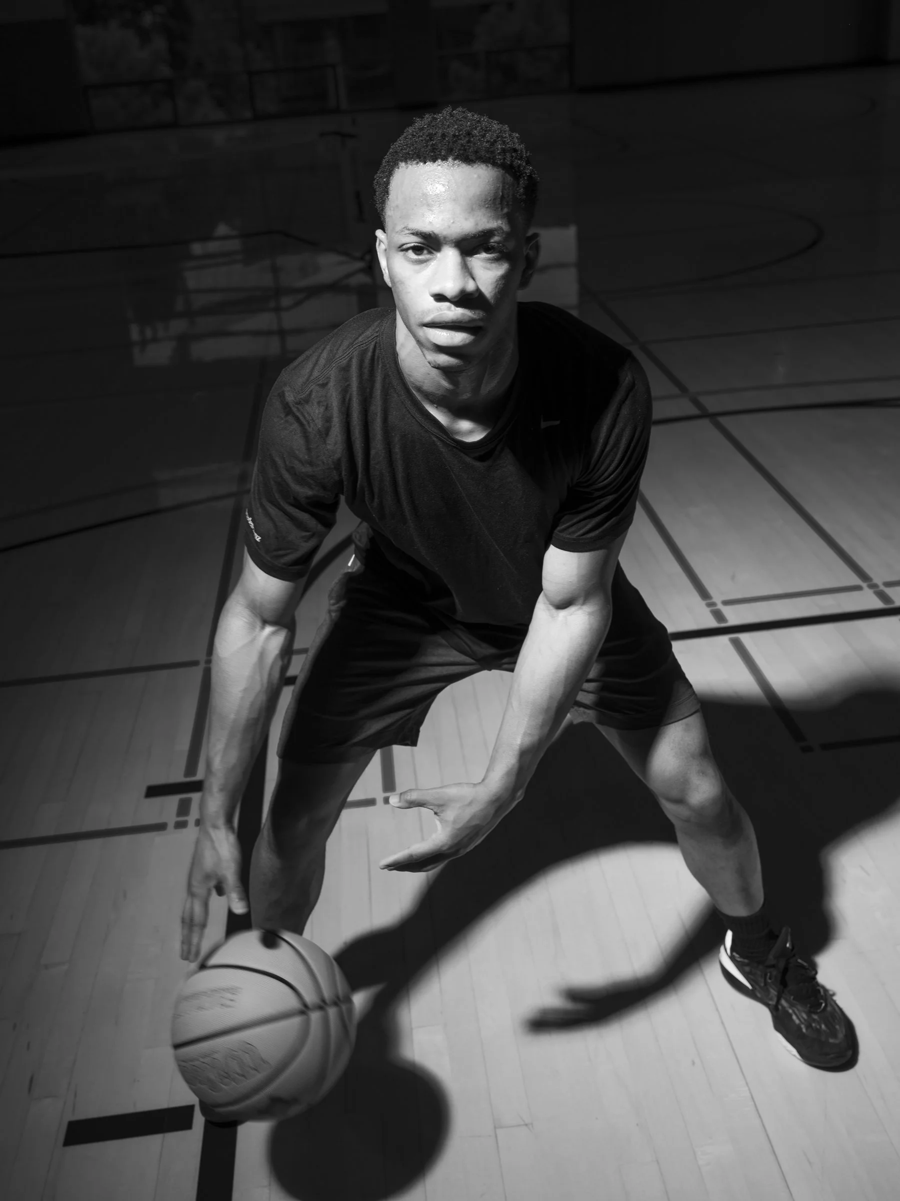 A young male basketball player is crouching on an indoor basketball court, holding a basketball with both hands. The image is in black and white with a spotlight effect highlighting his face and upper body.
