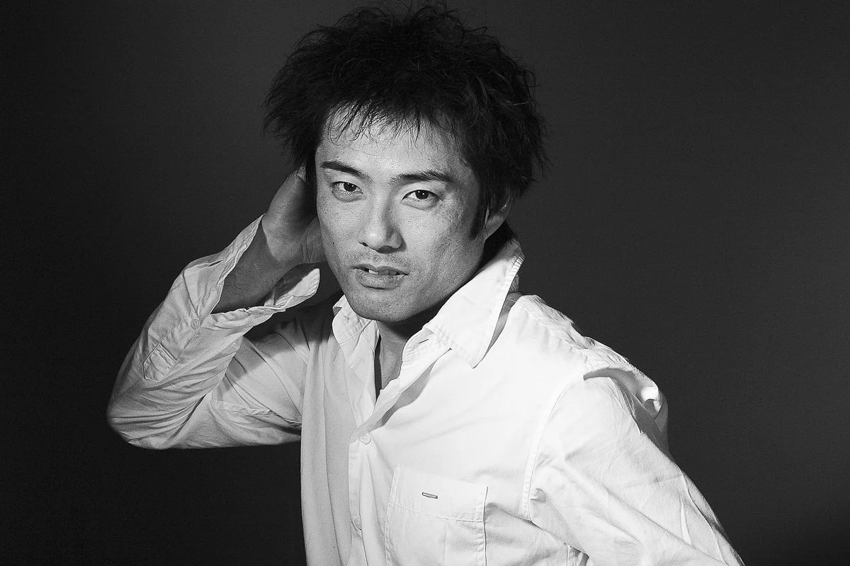 Black and white portrait of a man with tousled hair, wearing a white shirt, posing with a hand behind his head against a dark background.