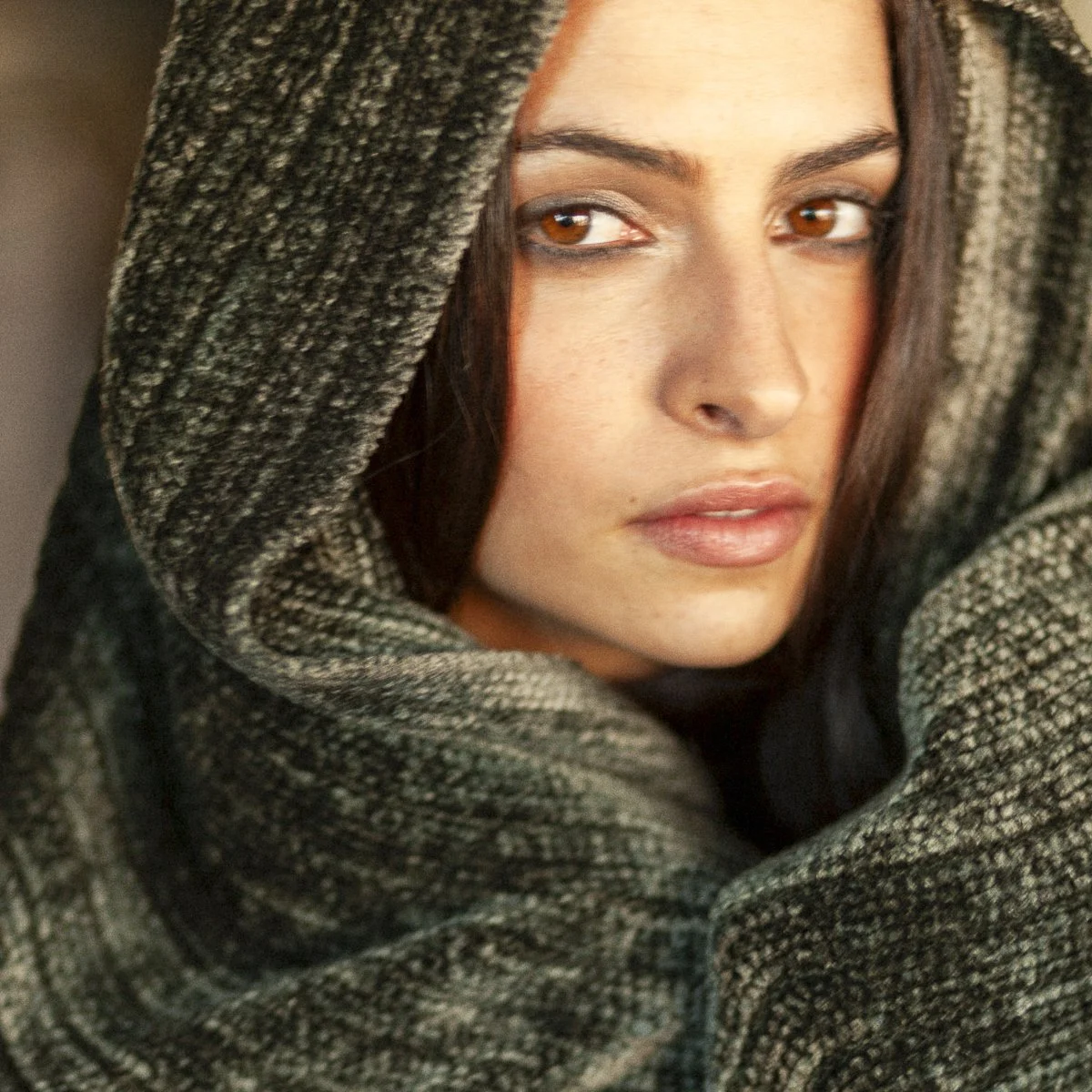 A woman with brown hair and fair skin wearing a gray-patterned hooded garment, gazing at the camera with a neutral expression.