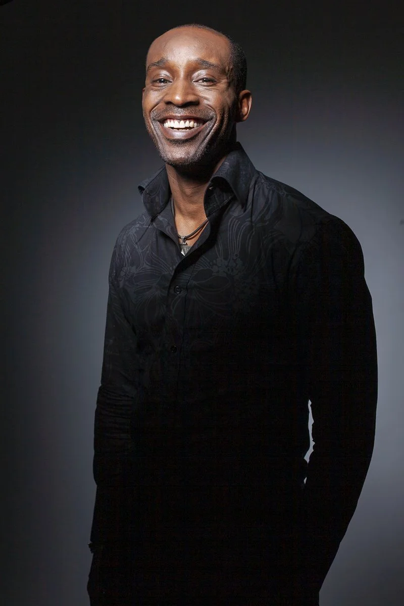 A smiling African American man in a black shirt with a dark background.