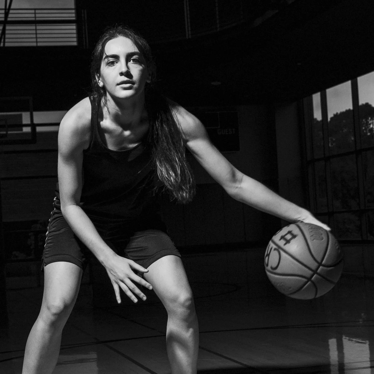 A young woman playing basketball on an indoor court, dribbling the ball with her right hand, wearing a tank top and shorts.