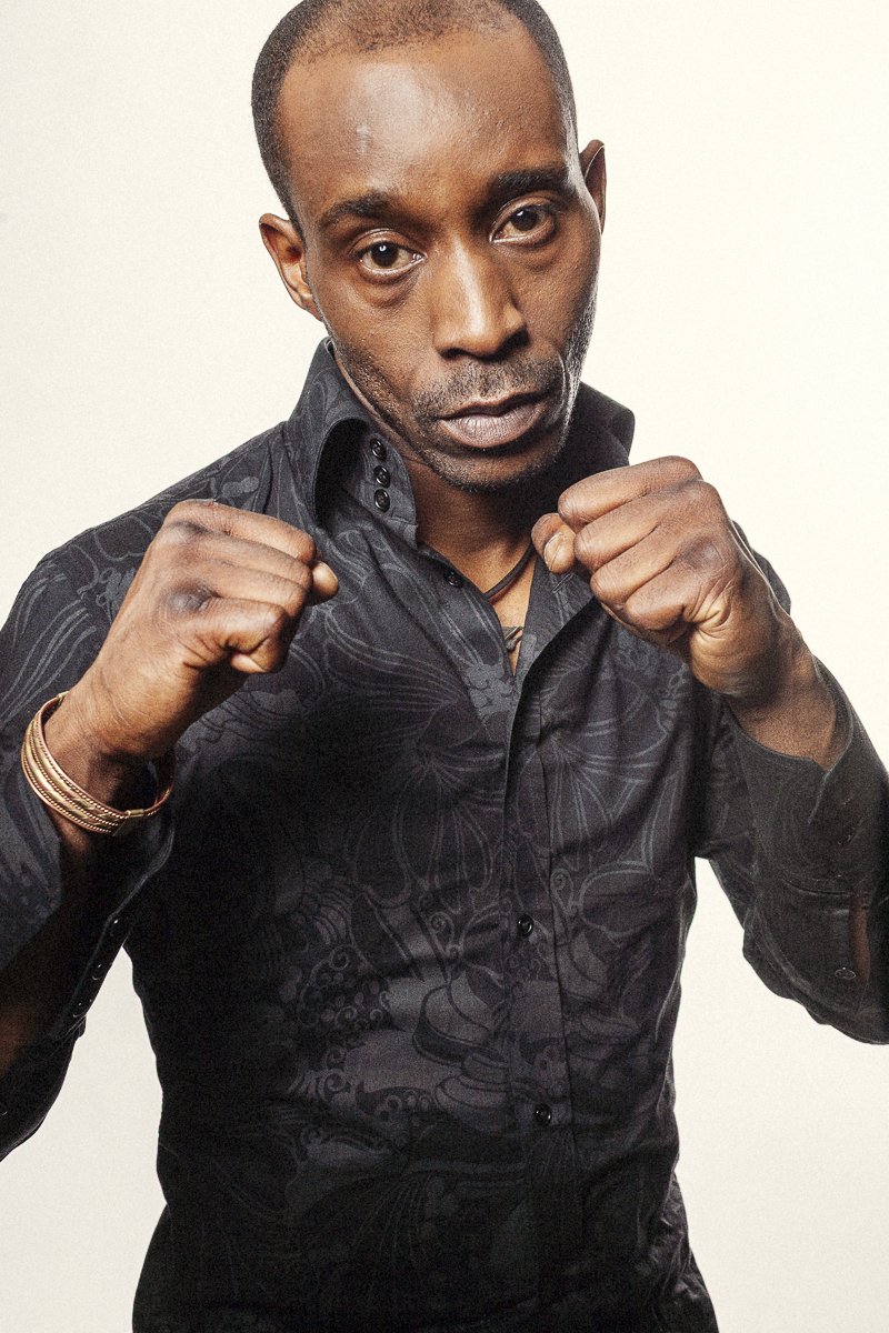 A man with a serious expression, wearing a black patterned shirt, holds his fists up in a boxing stance.