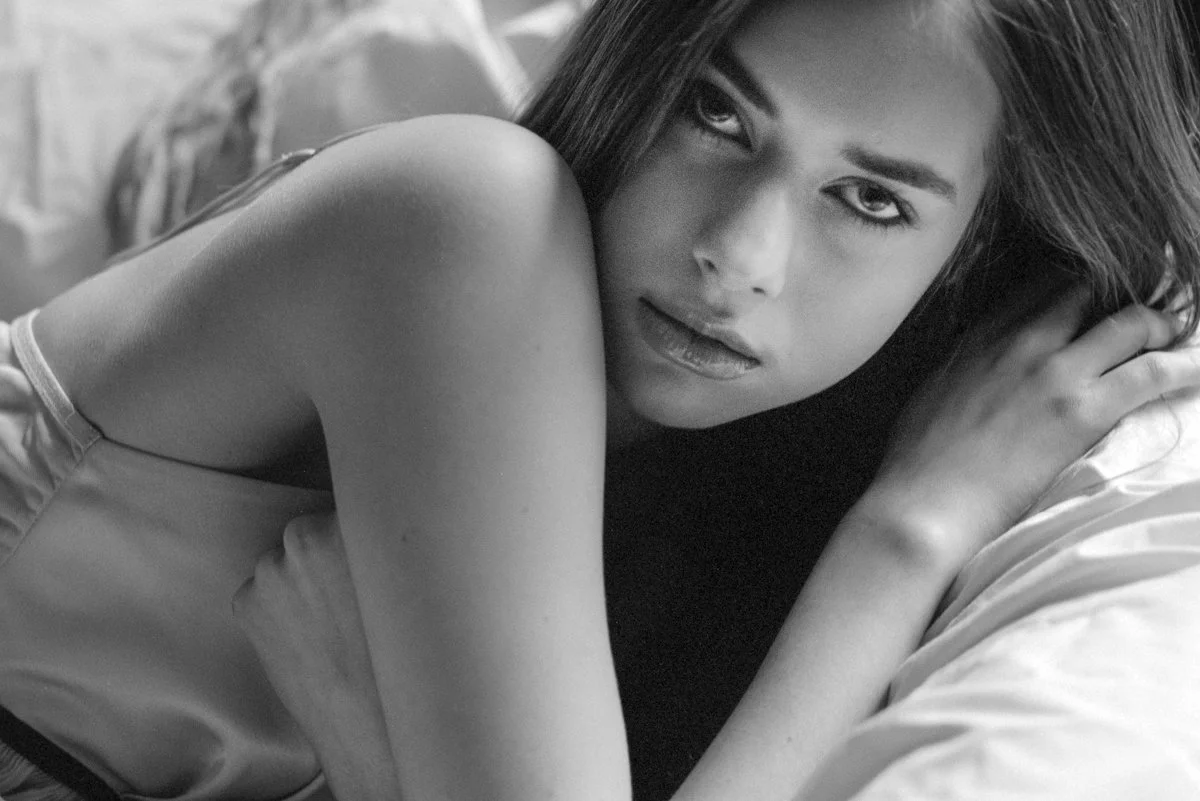 Close-up black and white photo of a woman with dark hair, lying on her side, looking directly at the camera.