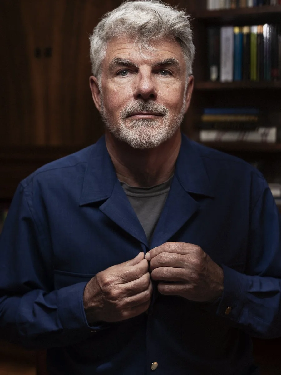 An elderly man with white hair and beard, wearing a navy blue shirt, is buttoning his shirt in front of shelves filled with books.