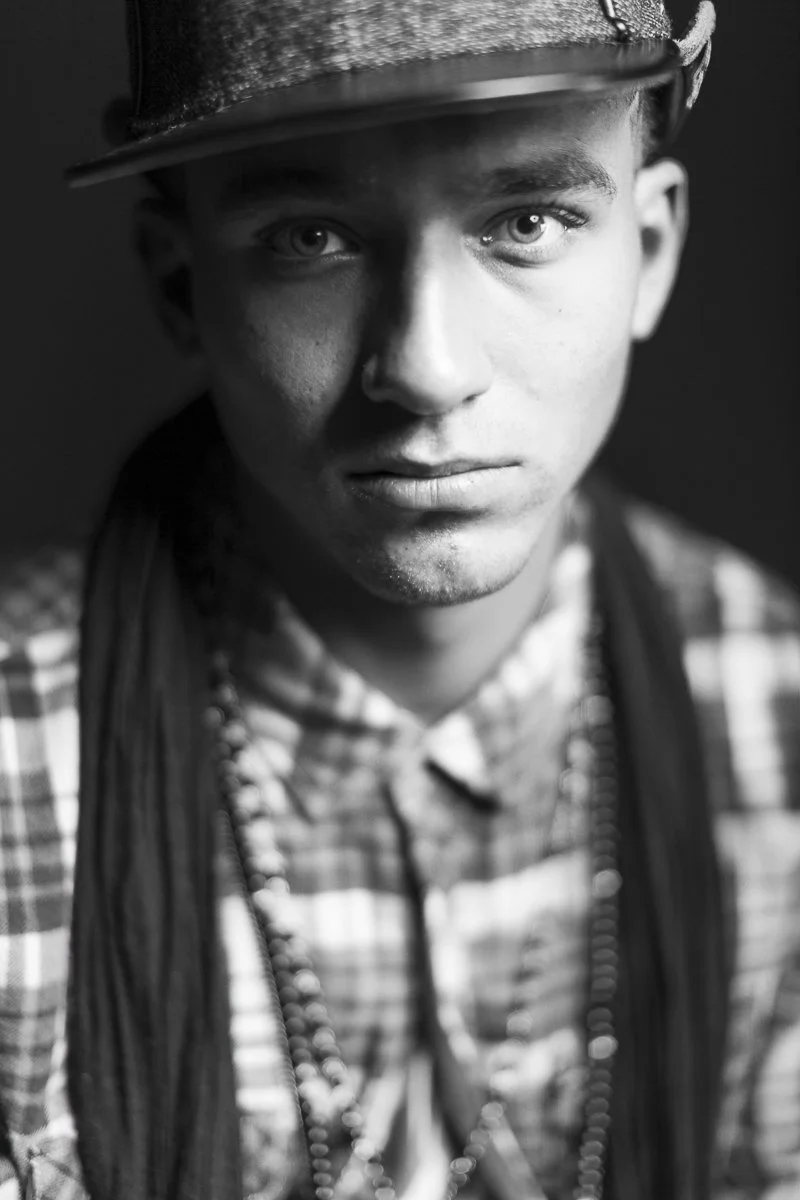 A black and white close-up portrait of a young man wearing a baseball cap tilted to the side, a checkered shirt, and layered necklaces. He has piercing eyes and a serious expression.