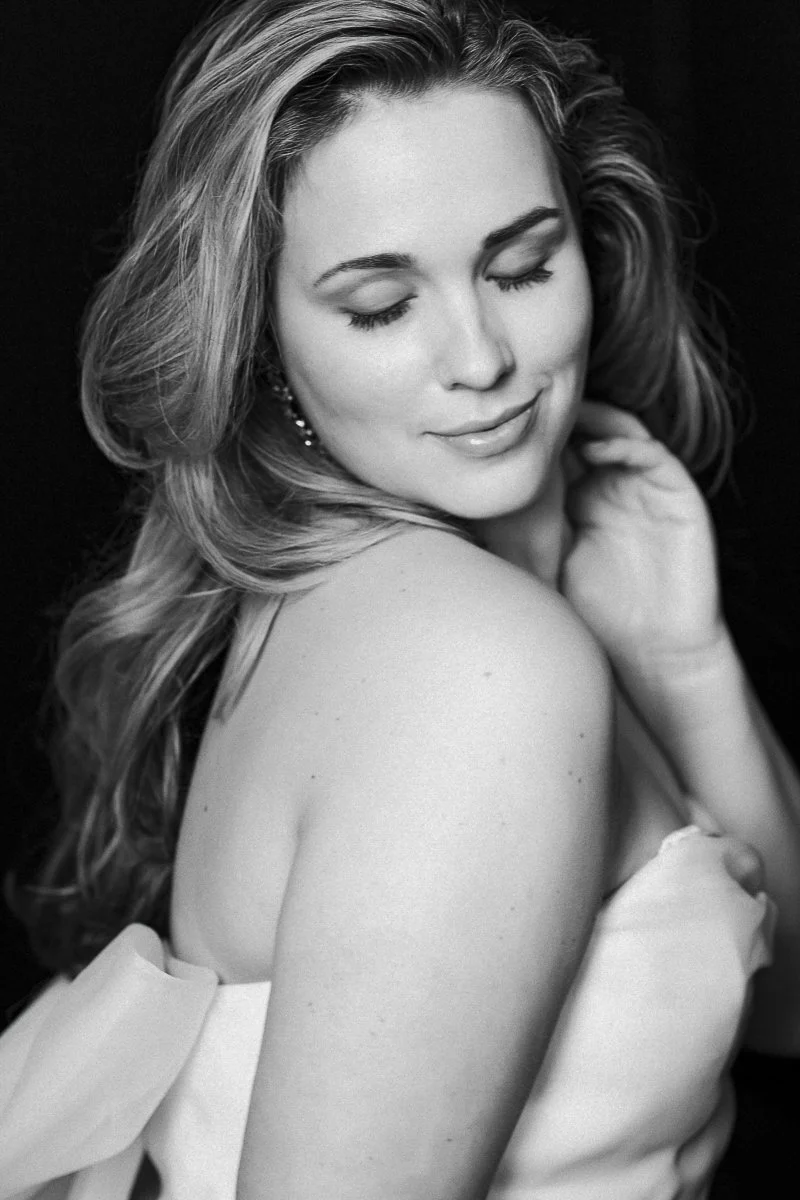 Black and white portrait of a smiling woman with closed eyes, long wavy hair, earrings, and a strapless dress, facing slightly to the right.
