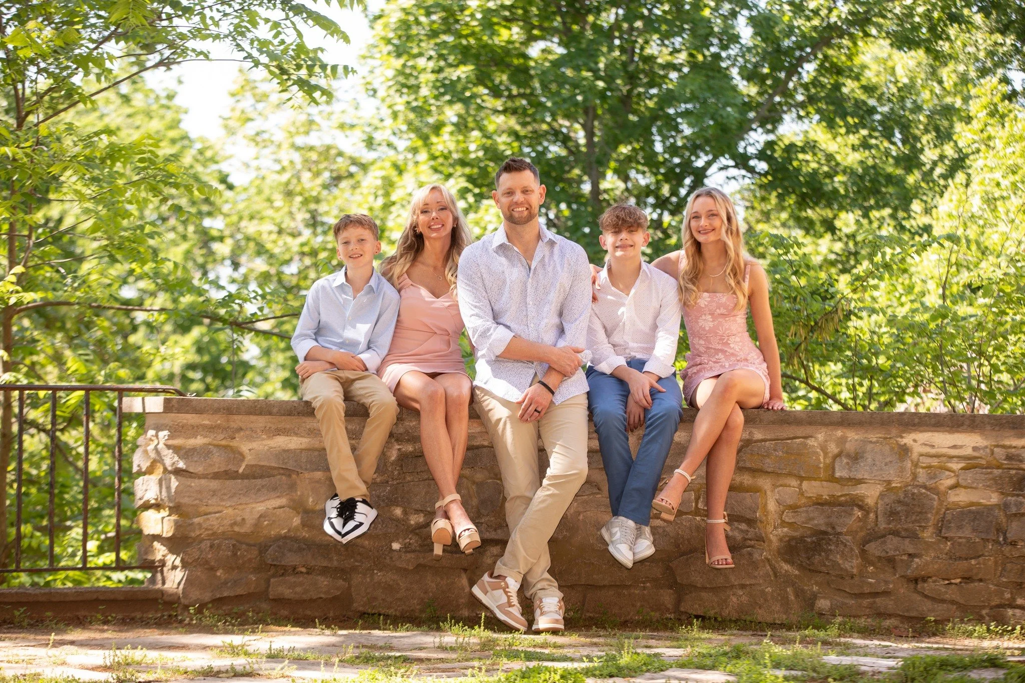 Family of five sitting on a stone wall outdoors surrounded by lush green trees. The family includes two children and three adults, all smiling at the camera, dressed in light-colored casual summer clothing.