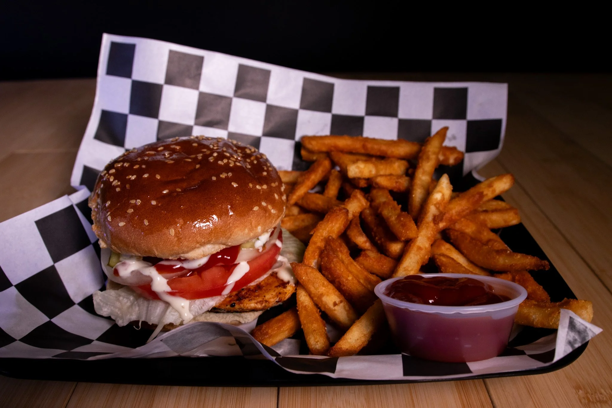 Burger with lettuce, tomato, mayonnaise, and sesame seed bun, served with French fries and a small cup of ketchup on a checkered paper-lined tray.