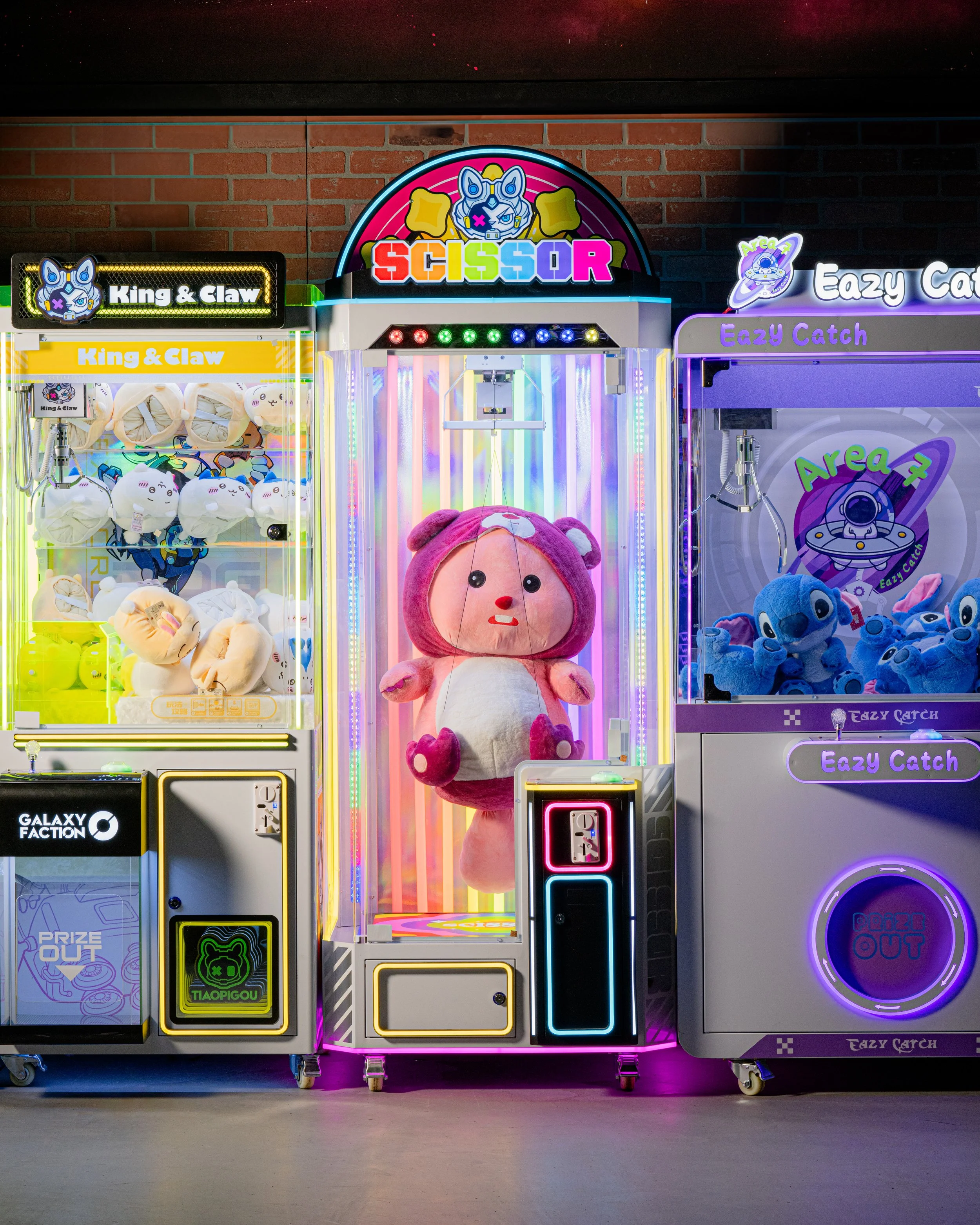 Colorful arcade claw machines with plush toys inside, featuring a large pink bear plush in the center and blue plush toys on the right, against a brick wall background.