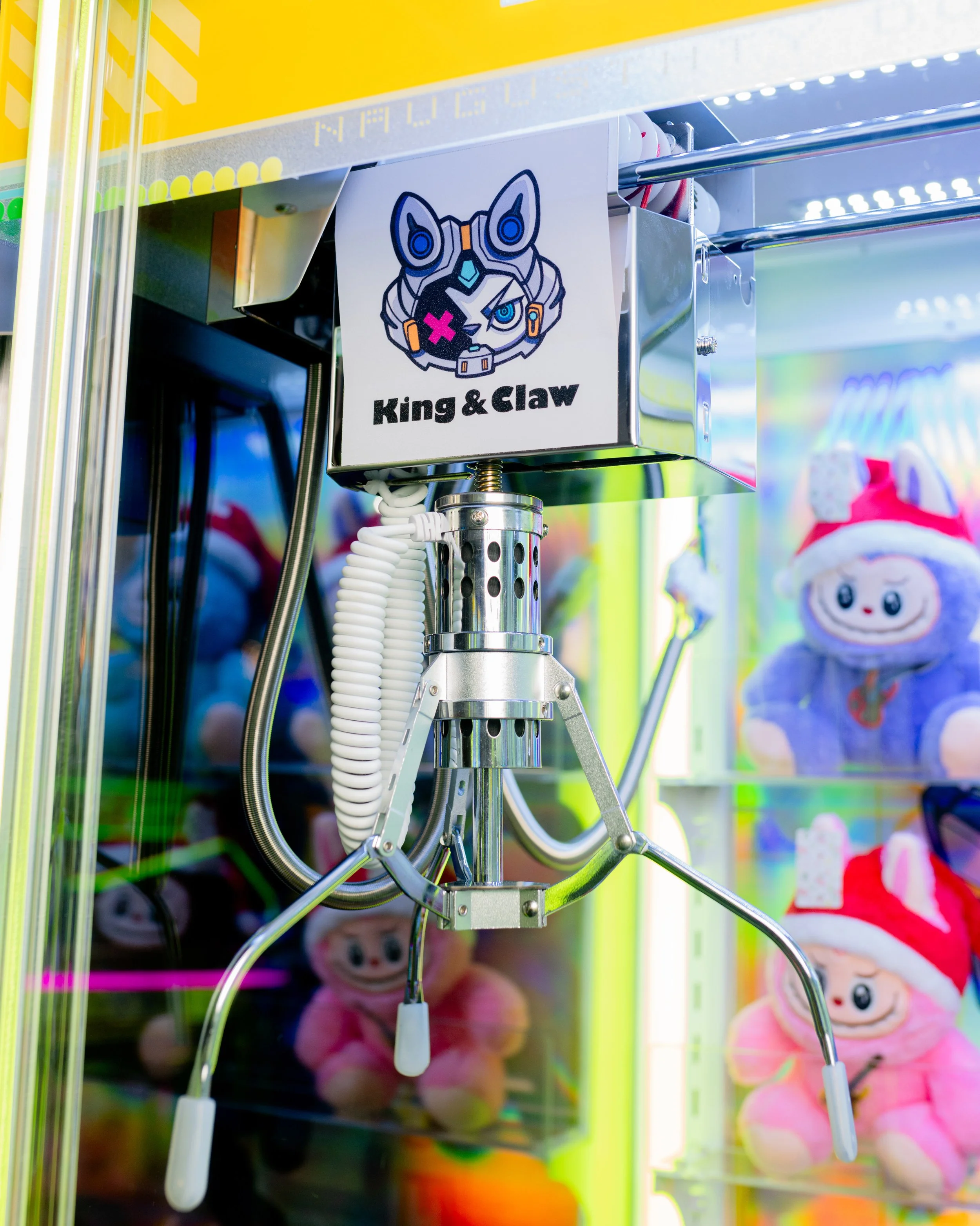 A claw machine with a sticker of a robotic animal face labeled 'King & Claw' and plush toys in the background.
