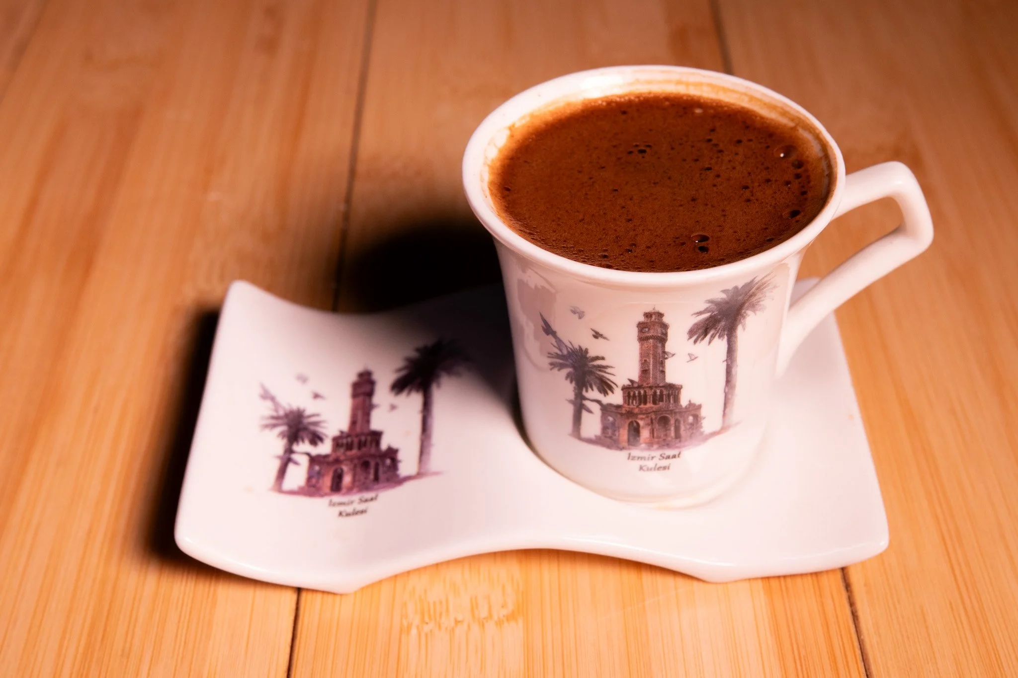 A white ceramic cup filled with Turkish coffee with frothy top, placed on a matching ceramic saucer with a scene of a tower and palm trees, on a wooden surface.