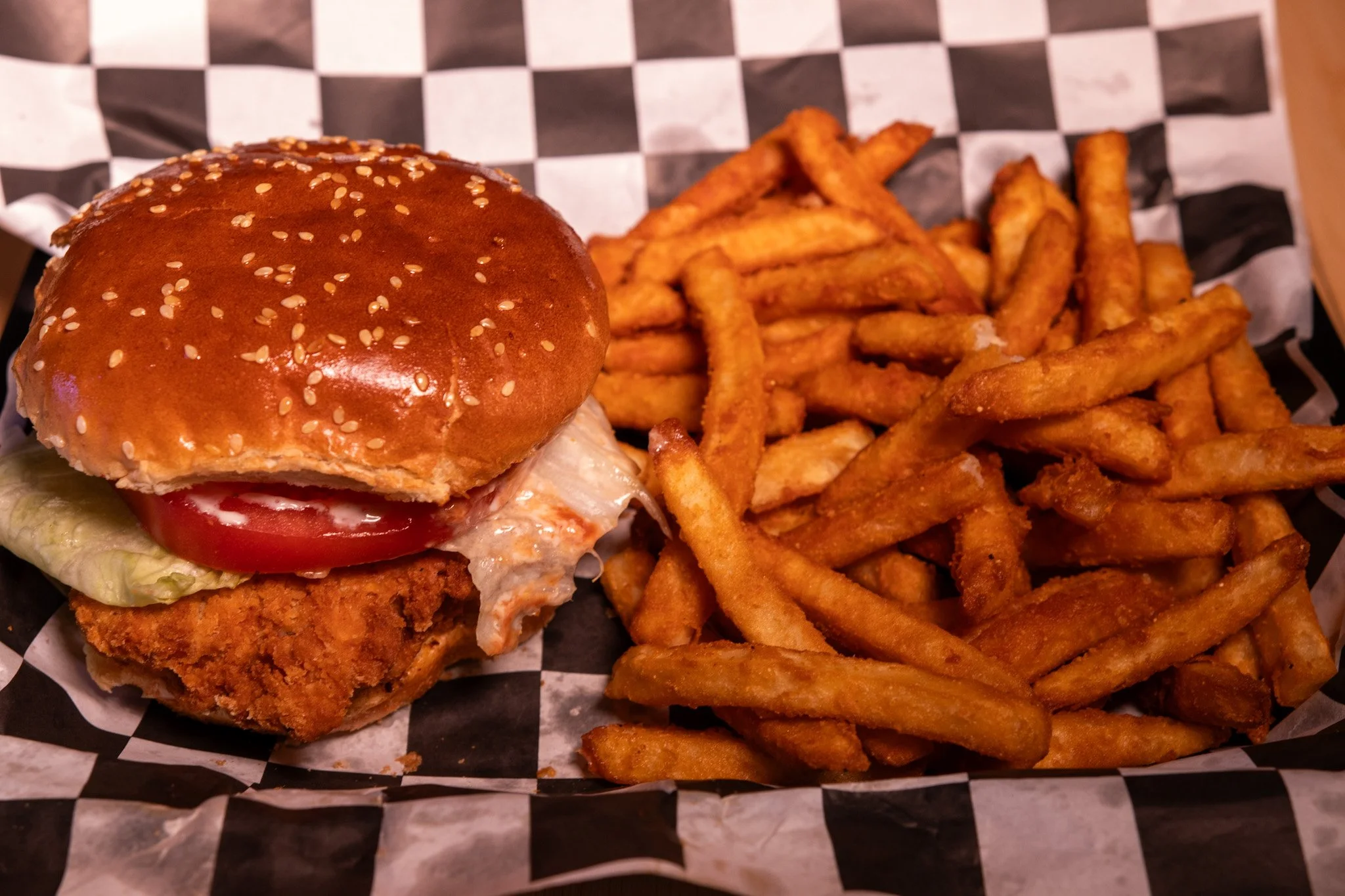Fried chicken sandwich with lettuce, tomato, and sauce in a sesame seed bun next to a side of French fries on checkered paper lining a basket.