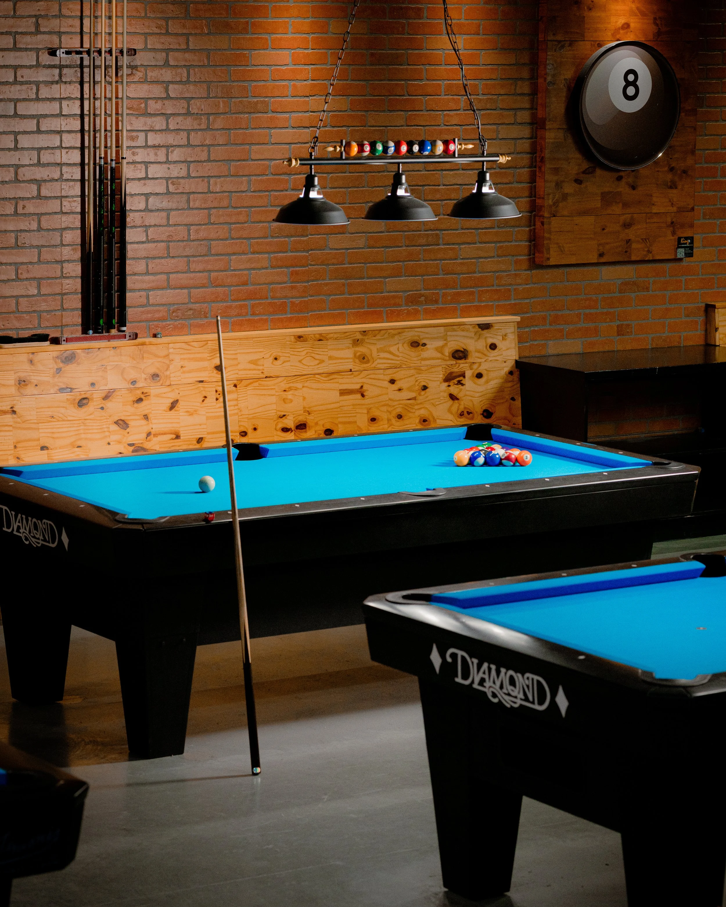 An indoor pool hall with two pool tables with blue felt and a brick wall. The table in focus has a cue stick and a rack of pool balls, with several balls racked and some scattered. Overhead, three black pool table lights hang, and a round eight ball clock is on the wall.