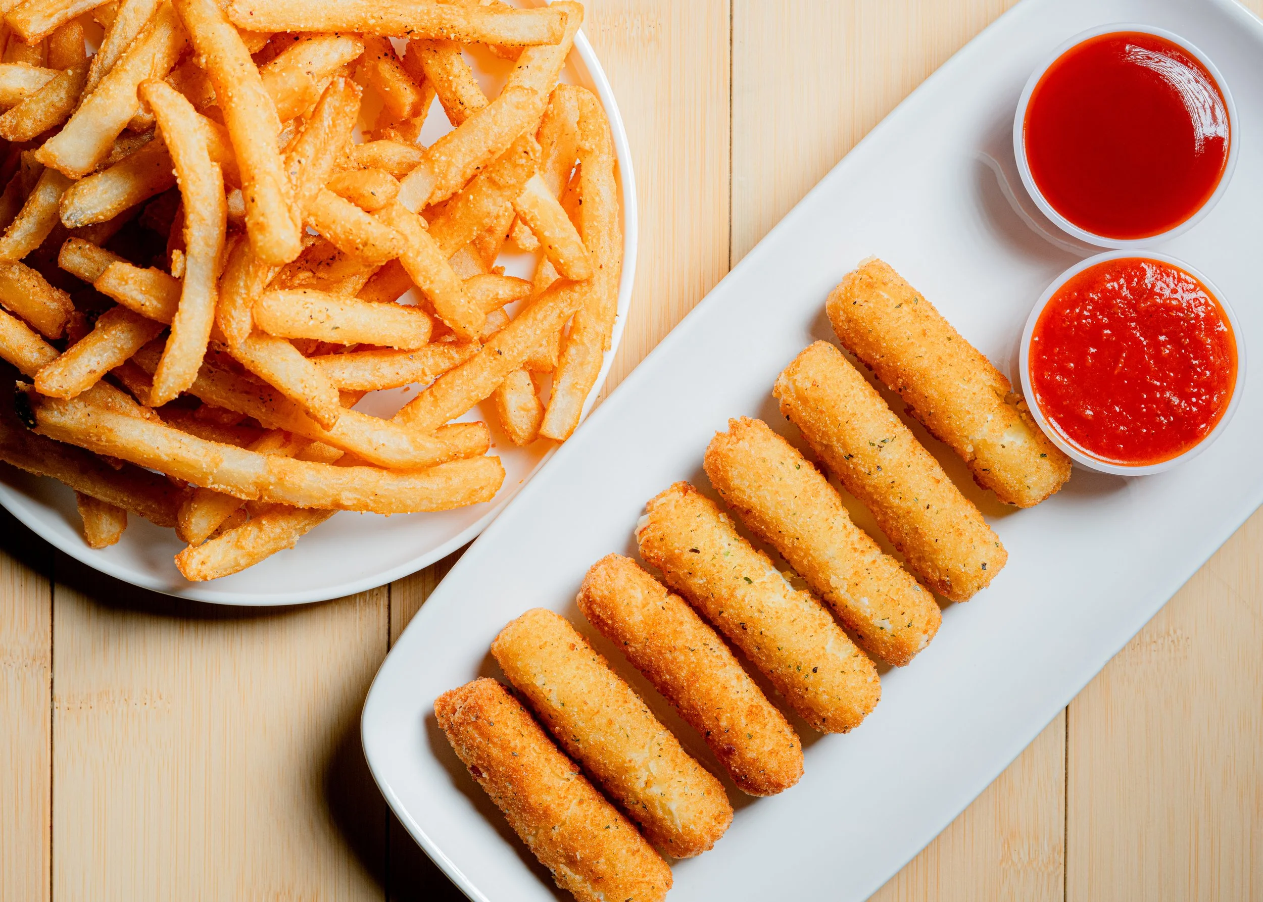 A plate of French fries, a row of mozzarella sticks with two cups of dipping sauces, one of marinara and the other of ketchup, on a wooden table.