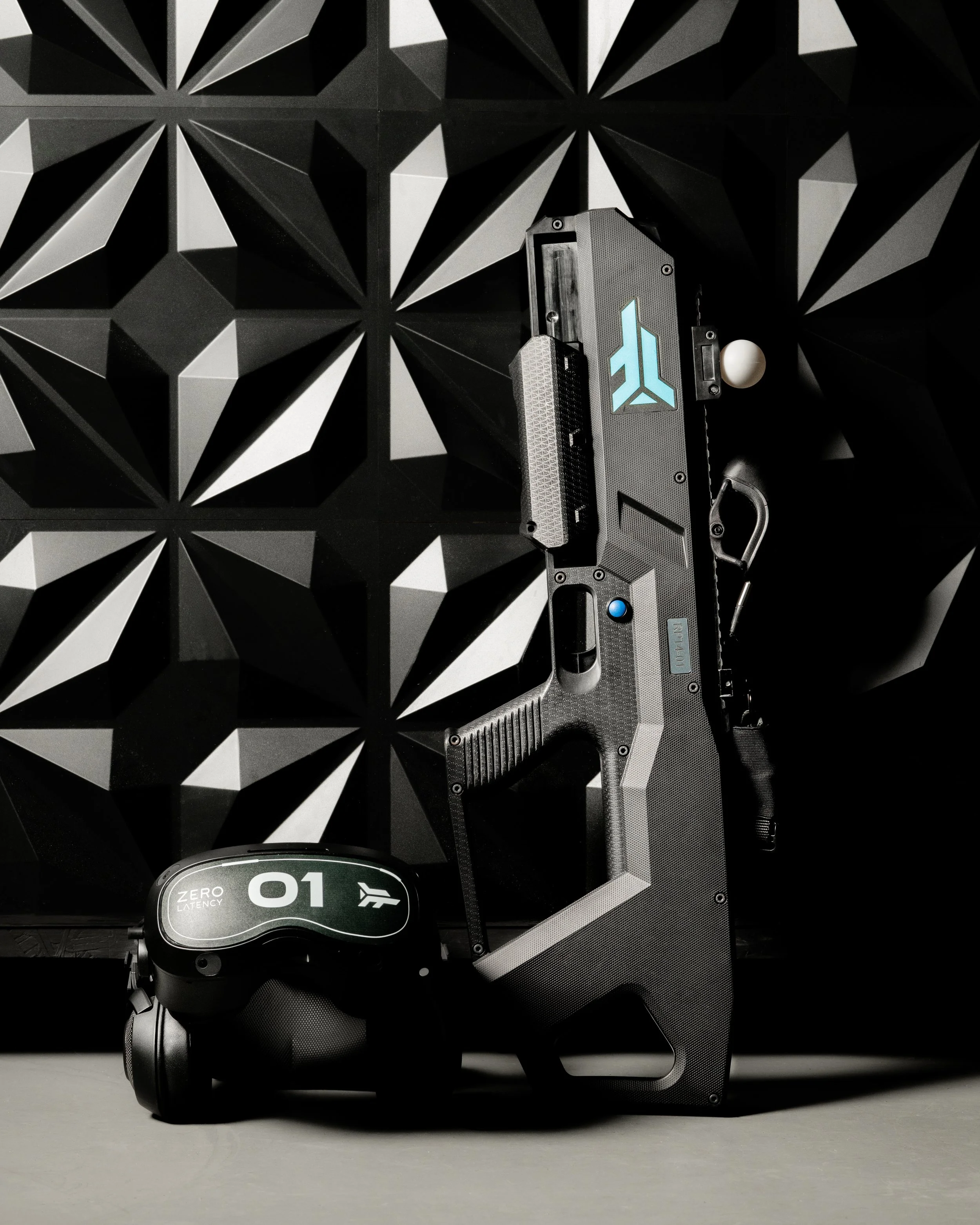 A black gaming gun controller with a blue button and a clip, placed in front of a geometric black and gray patterned wall, with a black virtual reality headset marked '01' and 'ZERO LATENCY' next to it.