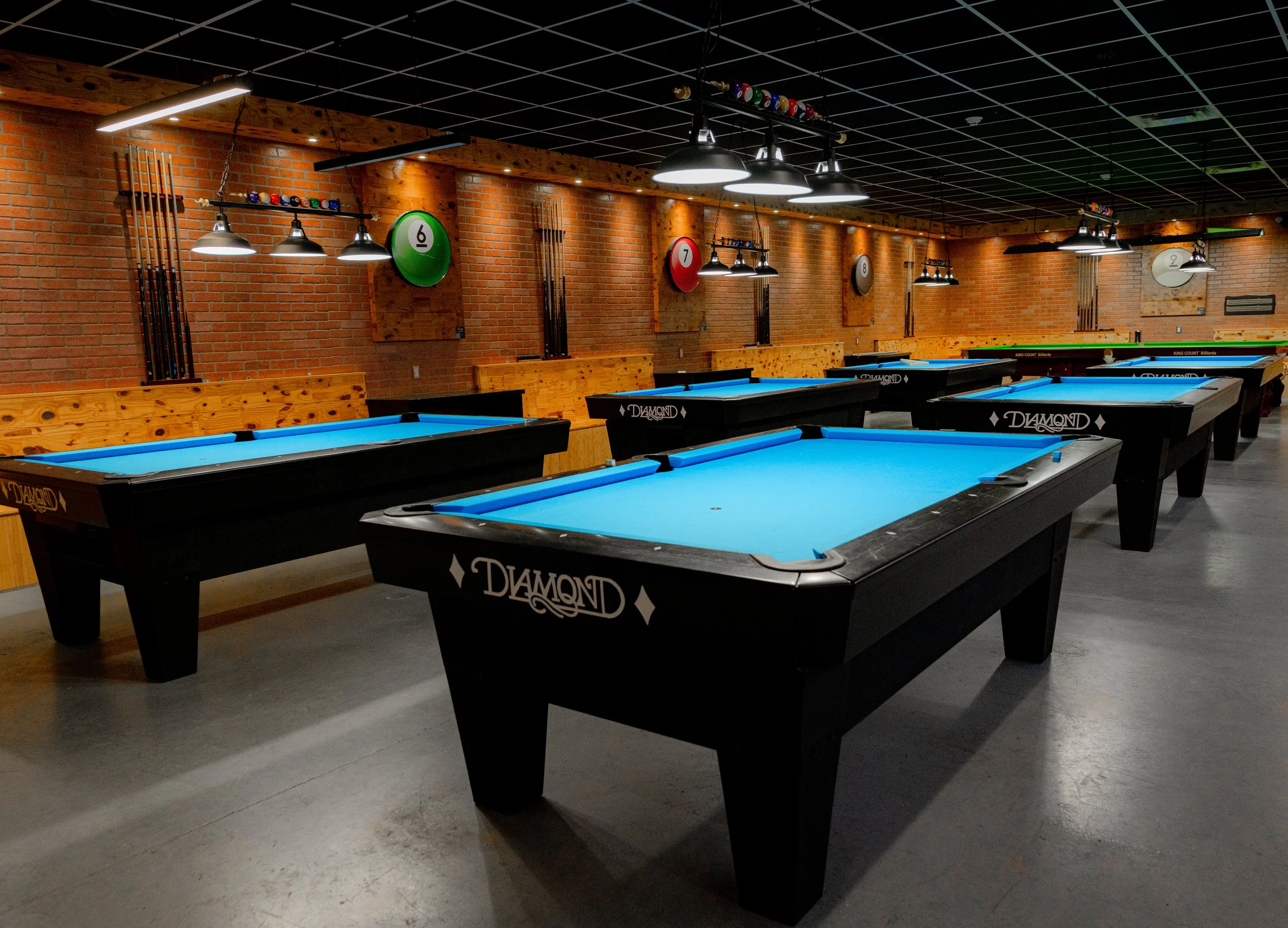 Indoor billiards room with multiple pool tables having blue felt, black frames with diamond logo, brick walls, wooden accents, pool cues mounted on wall, and overhead lighting.