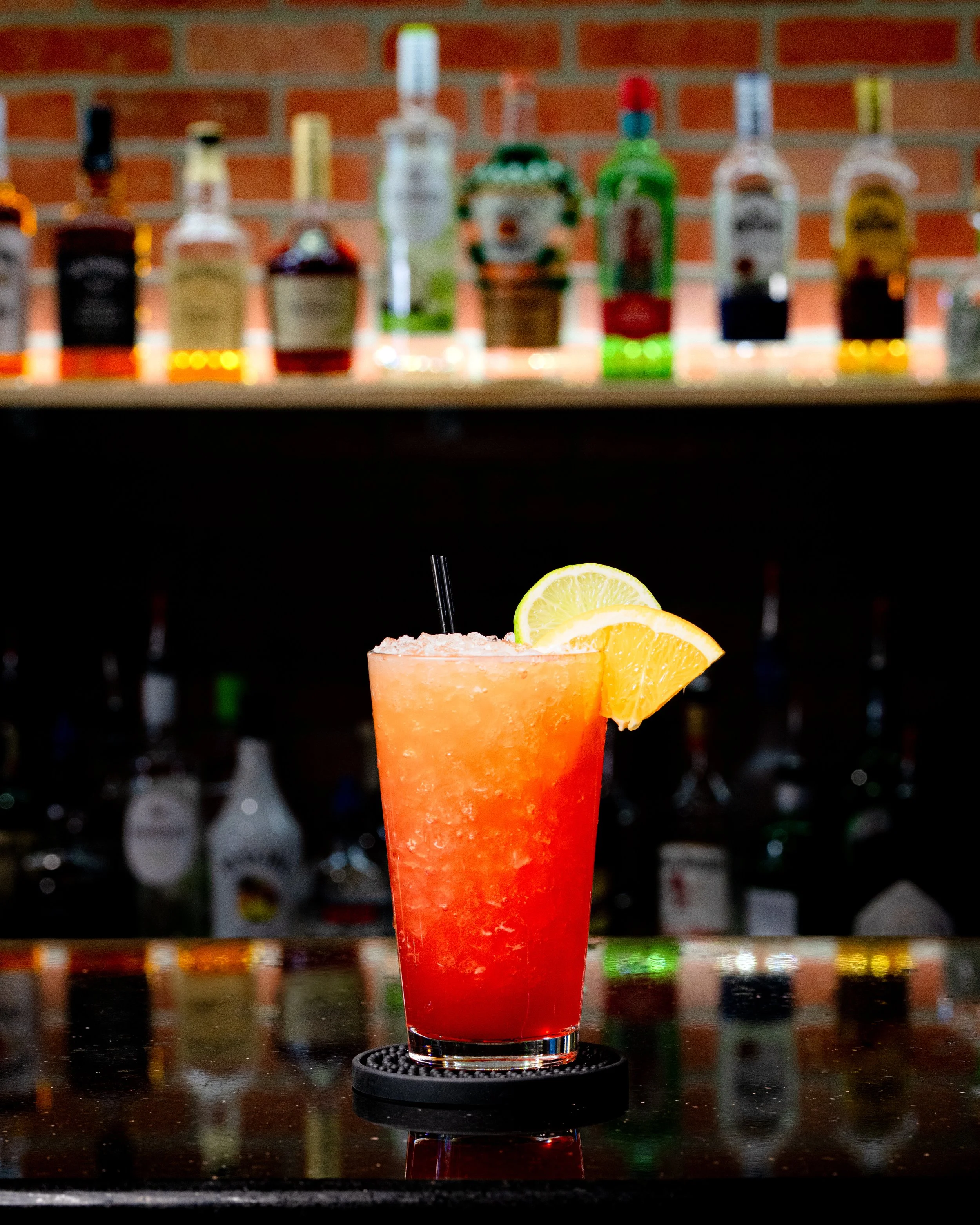 Bright red cocktail with lime and orange slices garnishing, placed on a black coaster on a reflective bar counter with blurred liquor bottles in the background.