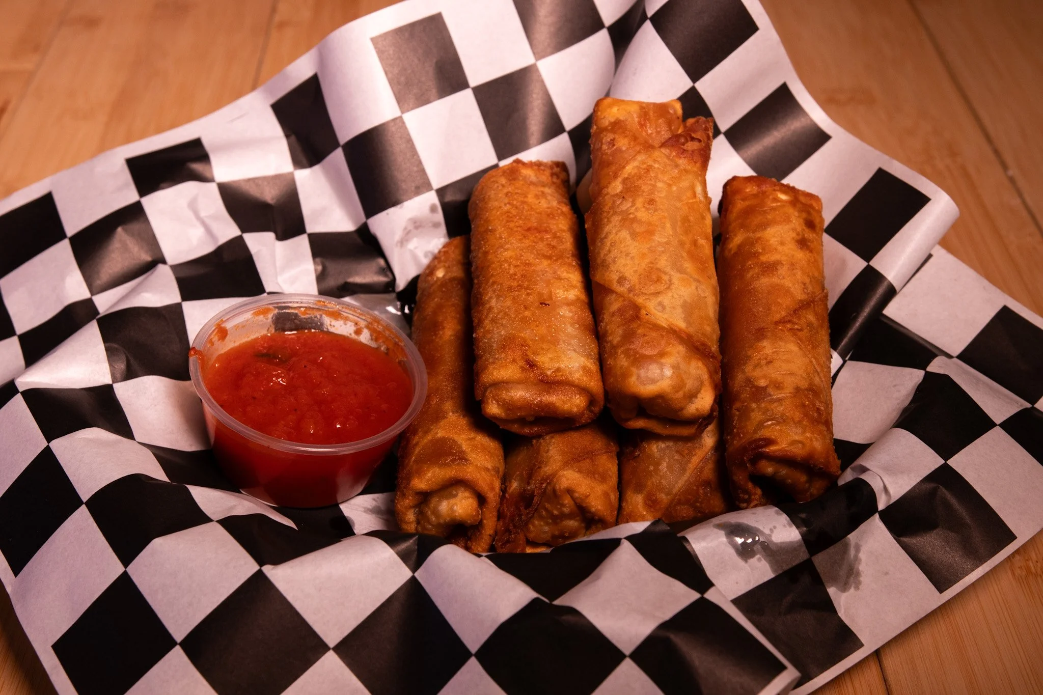 Six deep-fried egg rolls on checkered paper with a cup of red dipping sauce on the side.