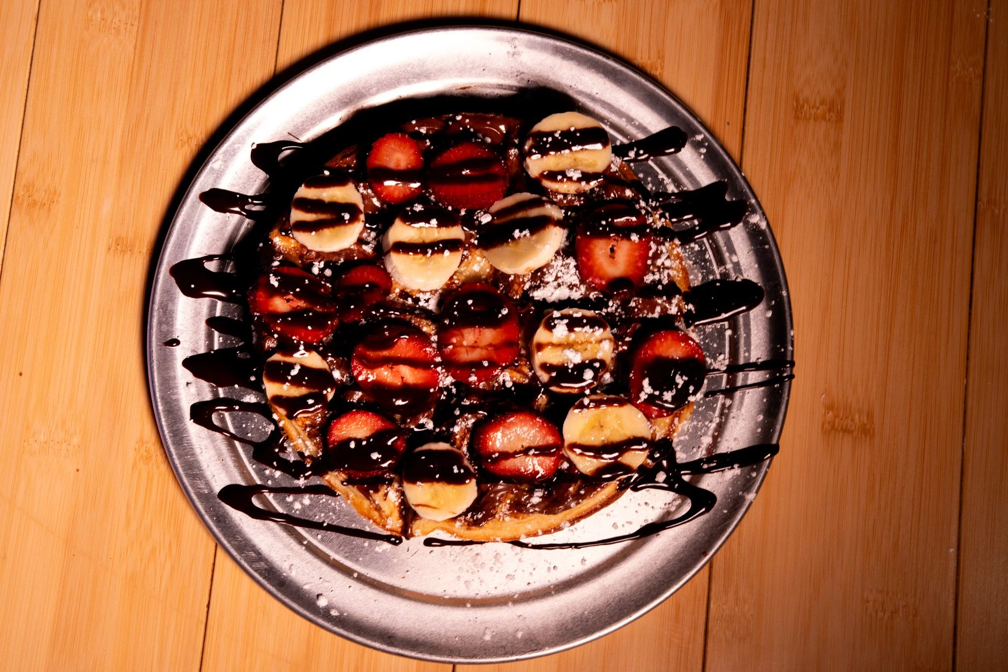 A plate of waffles topped with sliced bananas and strawberries, drizzled with chocolate syrup, dusted with powdered sugar, on a wooden table.