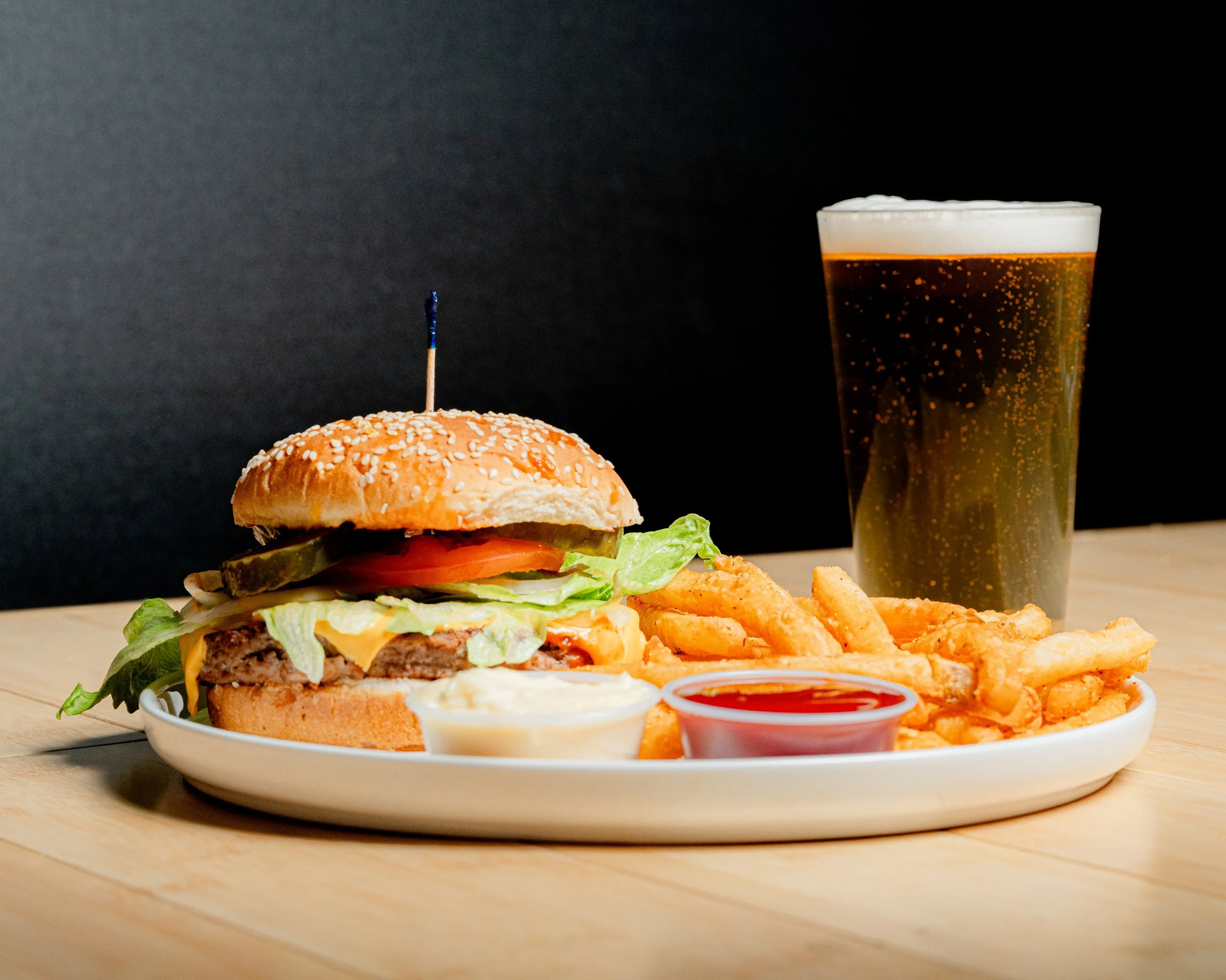A cheeseburger with lettuce, tomato, pickles, and pickled onions in a sesame seed bun with a toothpick, served with French fries, ketchup, and mayonnaise, and a glass of beer in the background on a wooden table against a black backdrop.