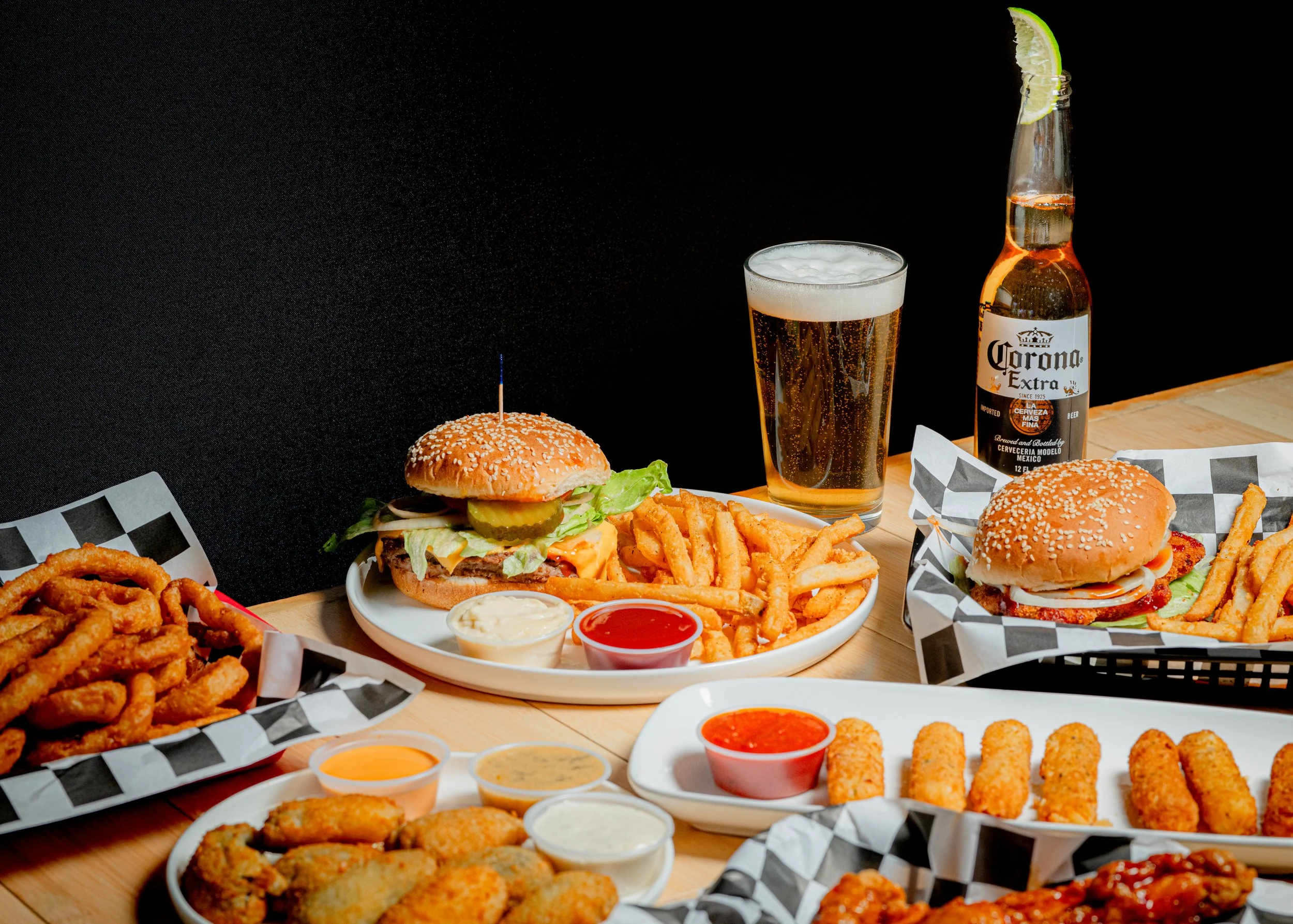 Various fast food items on a wooden table, including two cheeseburgers with fries, a basket of fried onion rings, chicken nuggets with dipping sauces, and mozzarella sticks with dipping sauce. There is also a glass of beer, a bottle of Corona beer with a lime wedge.