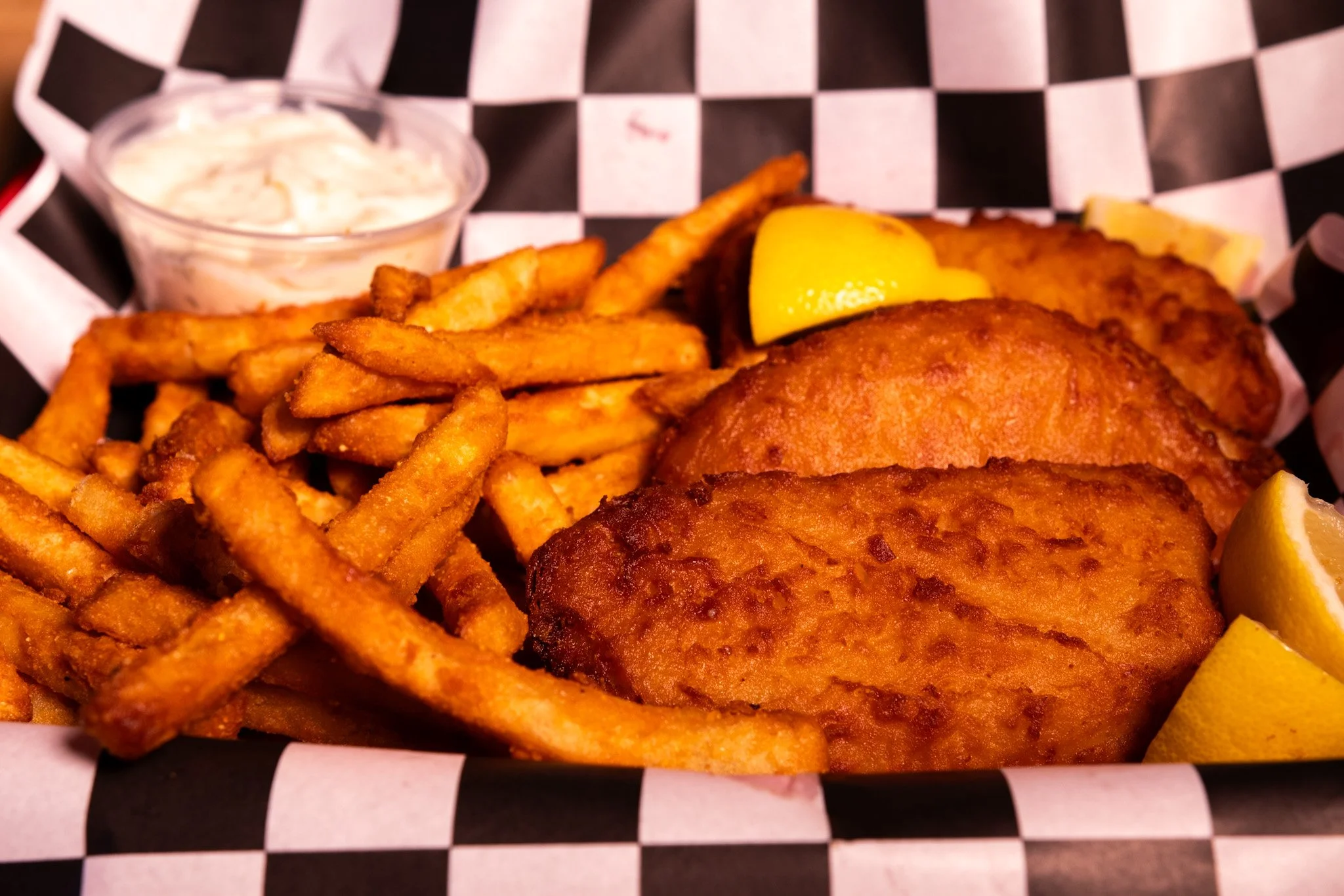 Fried fish fillets with lemon wedges, French fries, and a small cup of tartar sauce in a basket lined with black and white checkered paper.