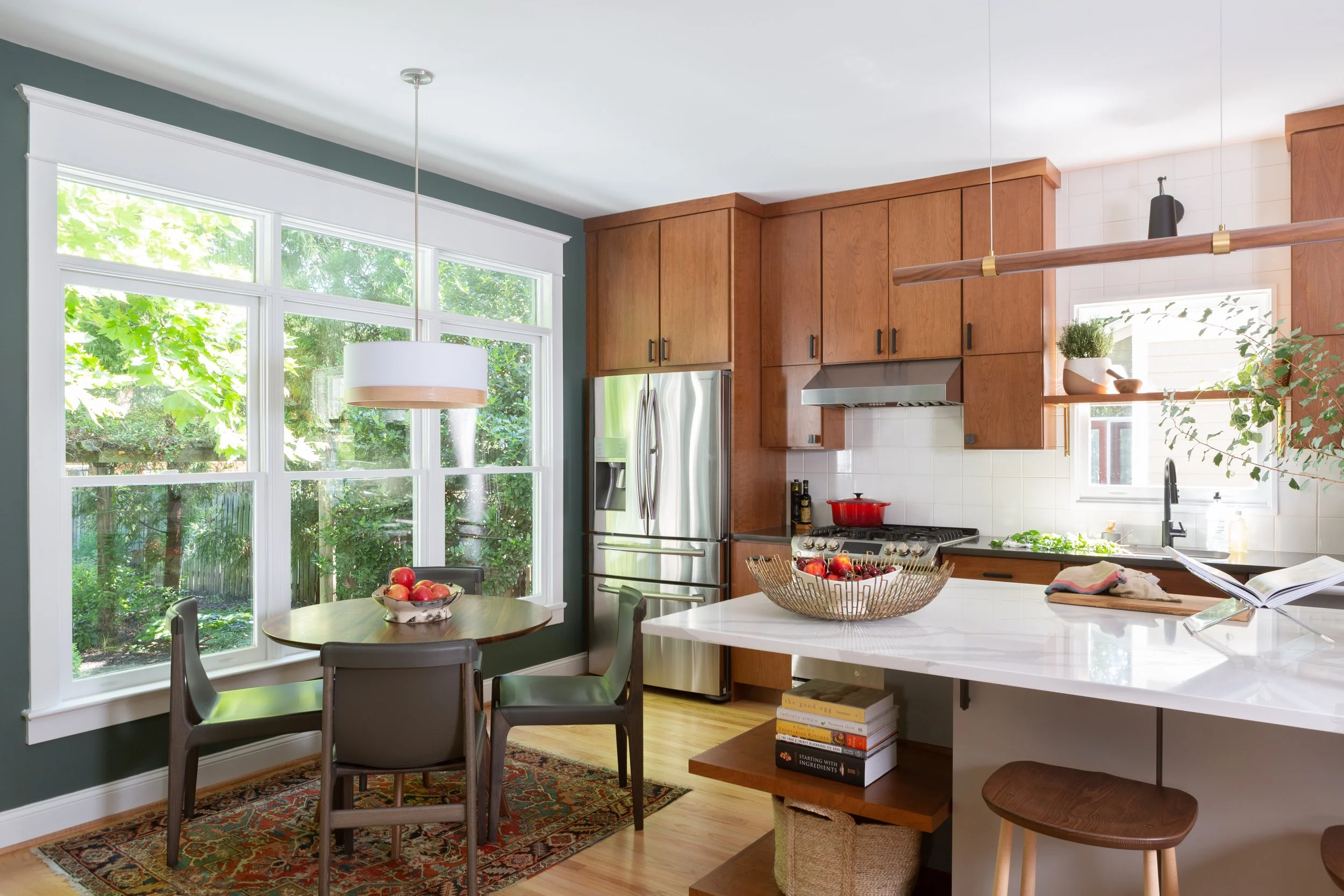 Kitchen with large window, wooden cabinets, stainless steel refrigerator, stove, white marble island, and dining table with four chairs.