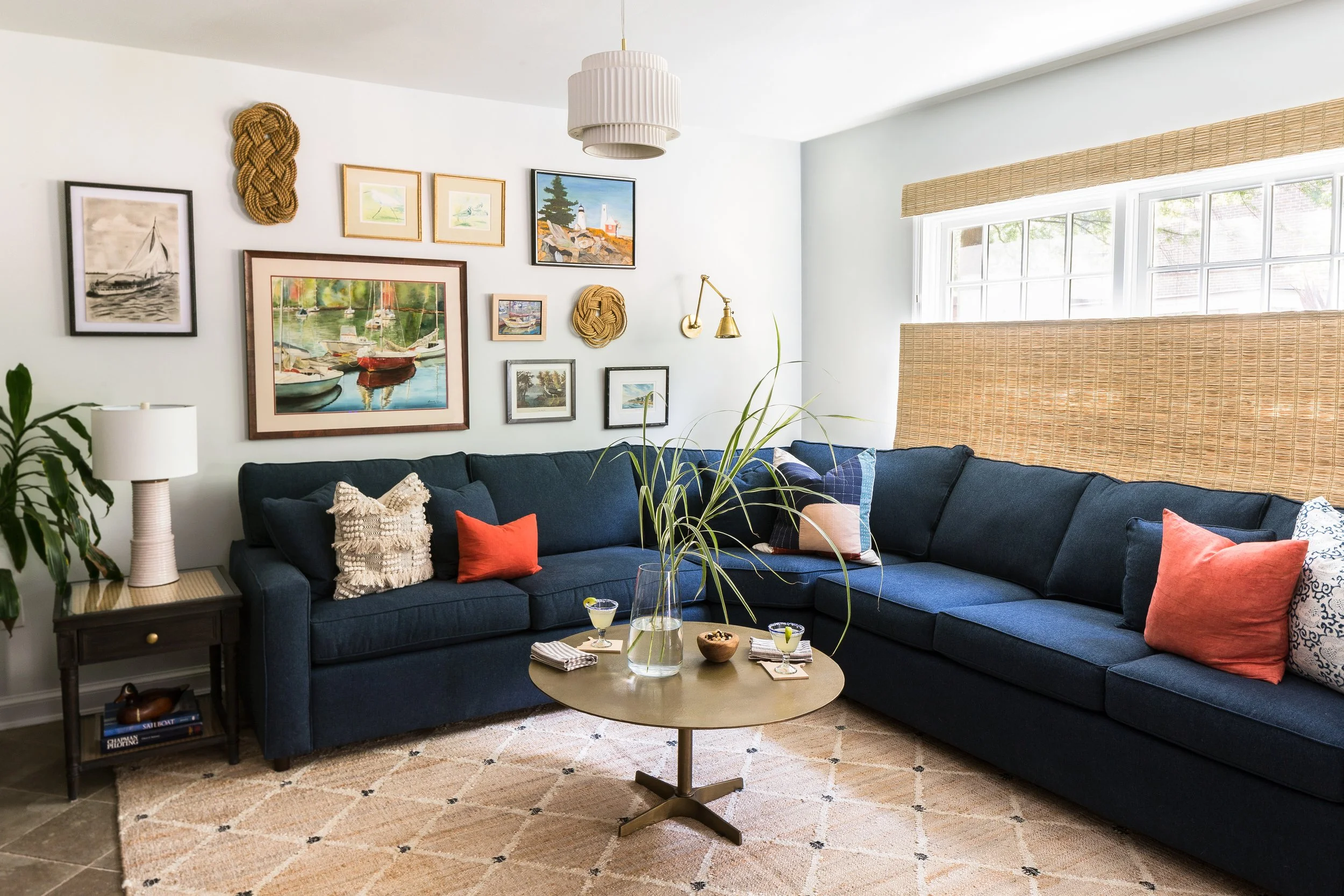 Living room with a navy blue sectional sofa, colorful throw pillows, a round coffee table with drinks, a side table with a lamp, a gallery wall with various framed artwork, a large window with woven shades, and a beige rug.