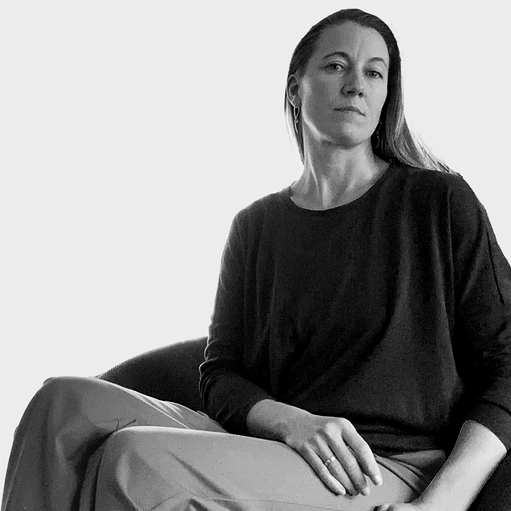 A woman with long hair and earrings, wearing a dark long-sleeve shirt, sitting on a chair against a plain background.