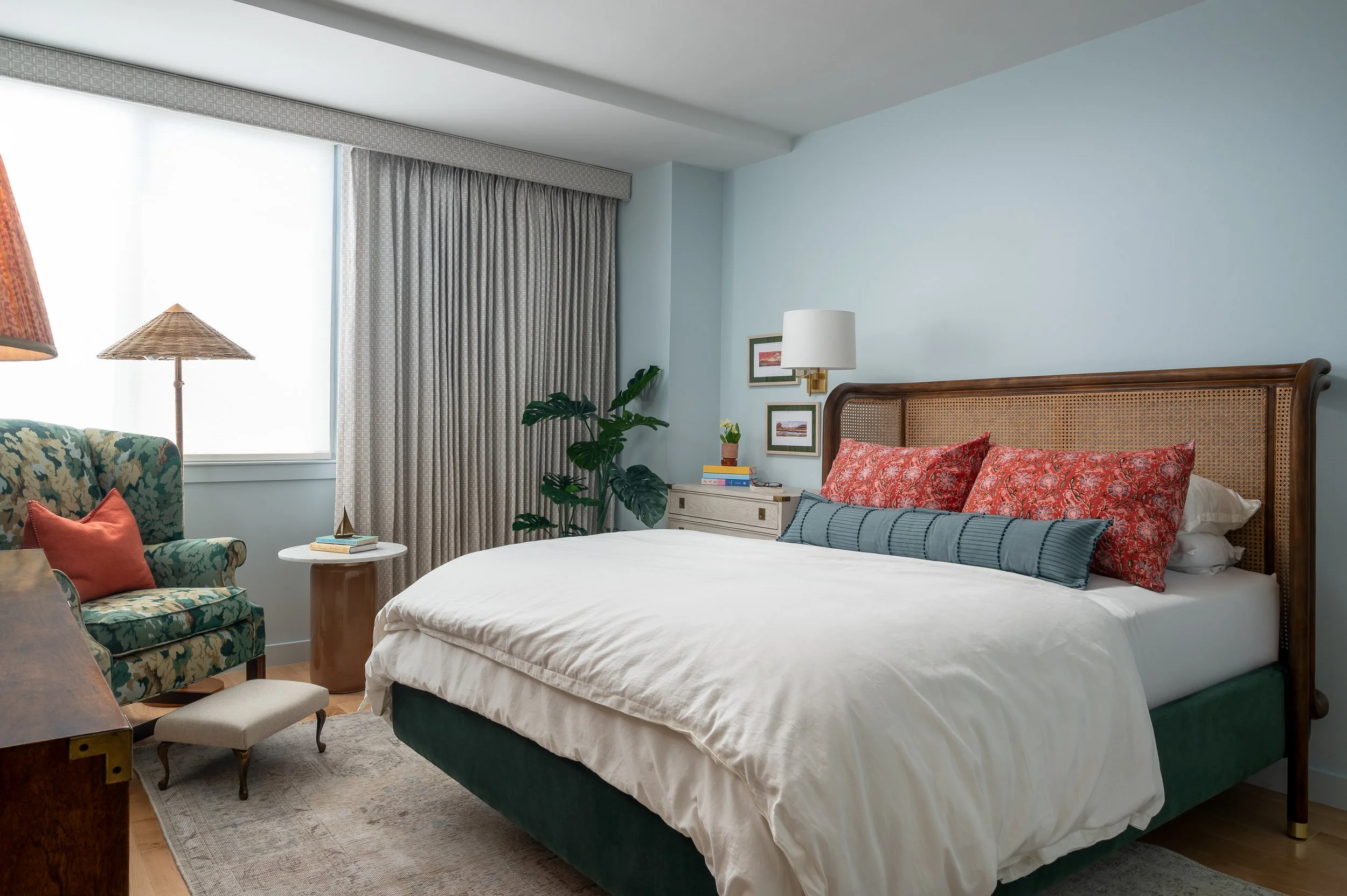 A bedroom with a large bed, bedside table, patterned armchair with a throw pillow, tall window with curtains, indoor plants, and wall art