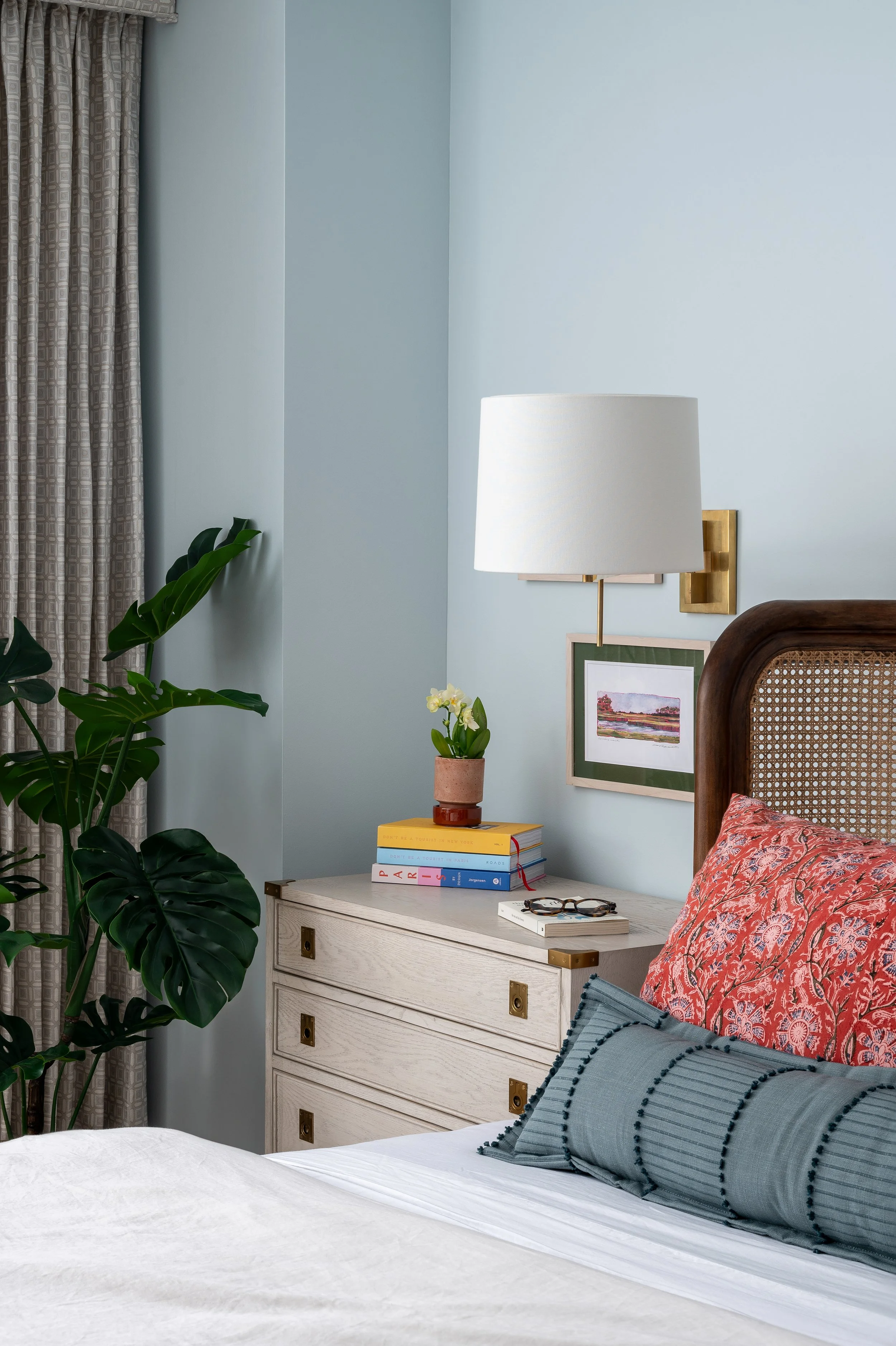 A bedroom corner with a white nightstand holding a potted plant, stacked books, and glasses, next to a bed with patterned pillows, a large leafy plant nearby, framed artwork and a white lampshade on the wall.