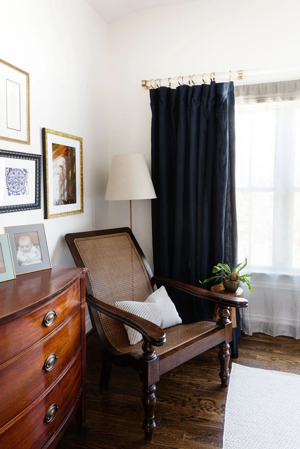 A cozy corner with a wooden armchair with a striped pillow, next to a small side table with a potted plant, a white lampshade, a window with dark and white curtains, and picture frames on the wall.