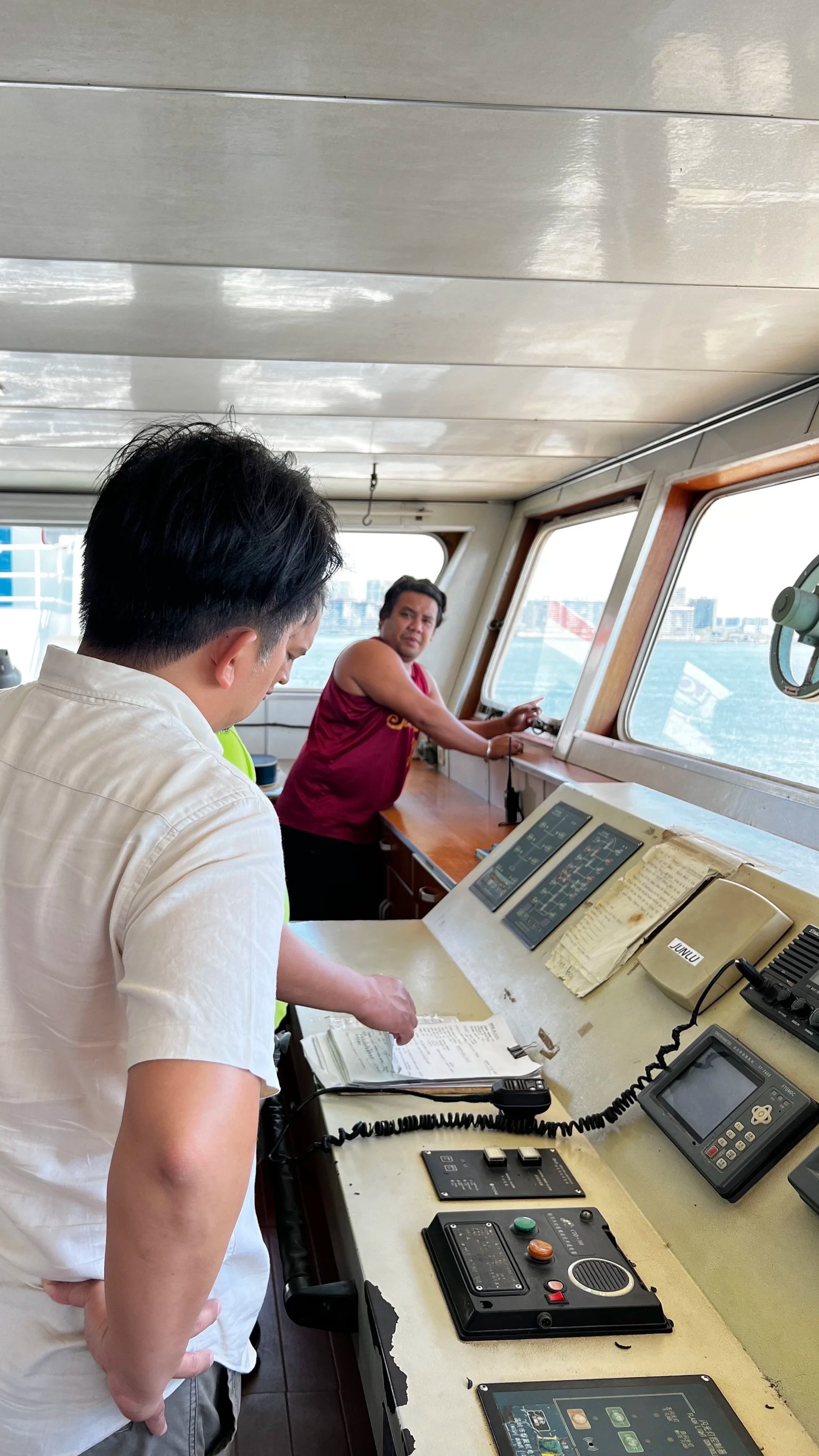 A man in a white shirt looking over papers at a ship's control station, with another man in a red sleeveless shirt standing near the windows, looking outward.