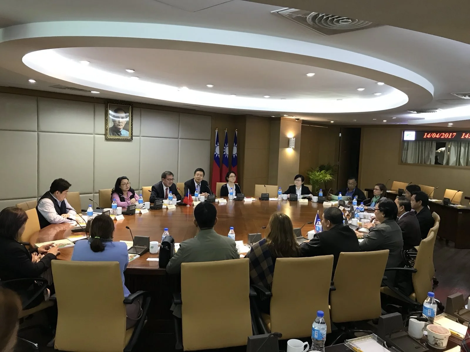 A large group of people seated around a U-shaped conference table in a formal meeting room, with flags and a portrait on the wall behind them.