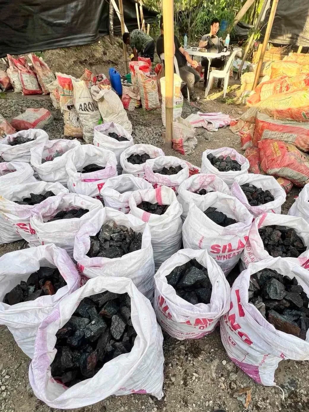 Bags of charcoal at an outdoor market stall with some people sitting and working in the background.