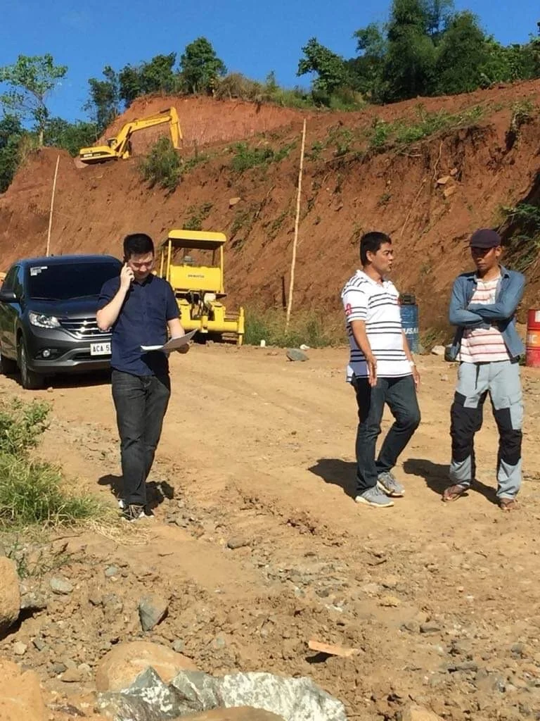 Three men on a construction site with excavation equipment; one is on the phone with documents, two are standing and talking, with a yellow excavator on a dirt hill in the background.