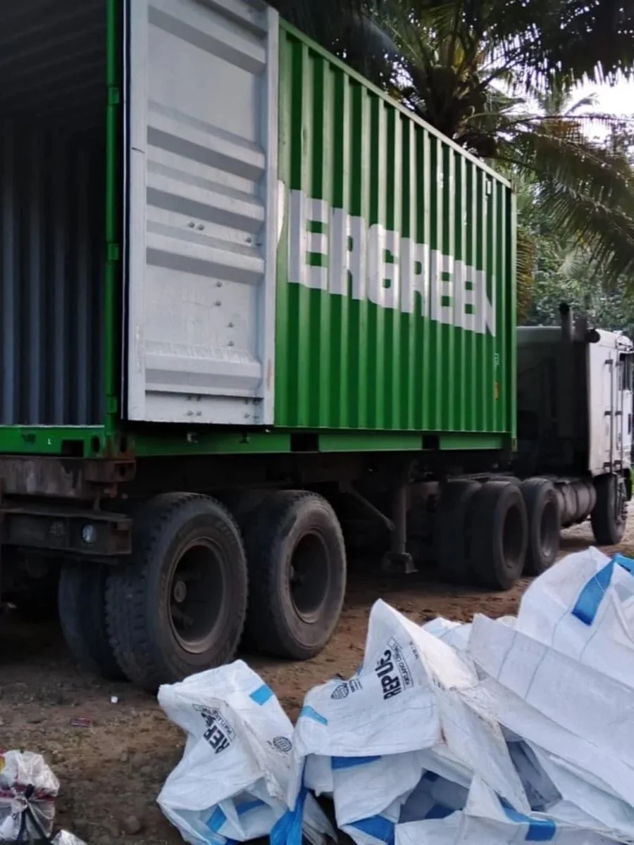 A large truck with a green container, which has white lettering on the side, parked on a dirt surface. There are white bags and debris on the ground in the foreground, and tall palm trees are visible in the background.