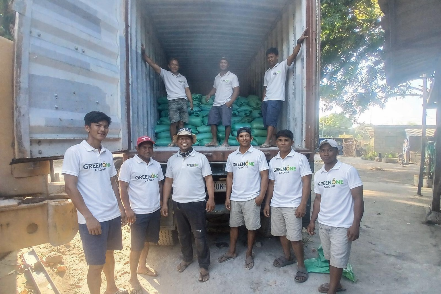 Group of men standing in front of a truck filled with green bags, with some people inside the truck. All are wearing white shirts with the GREEN ONE logo, suggesting they are part of a community or environmental group.