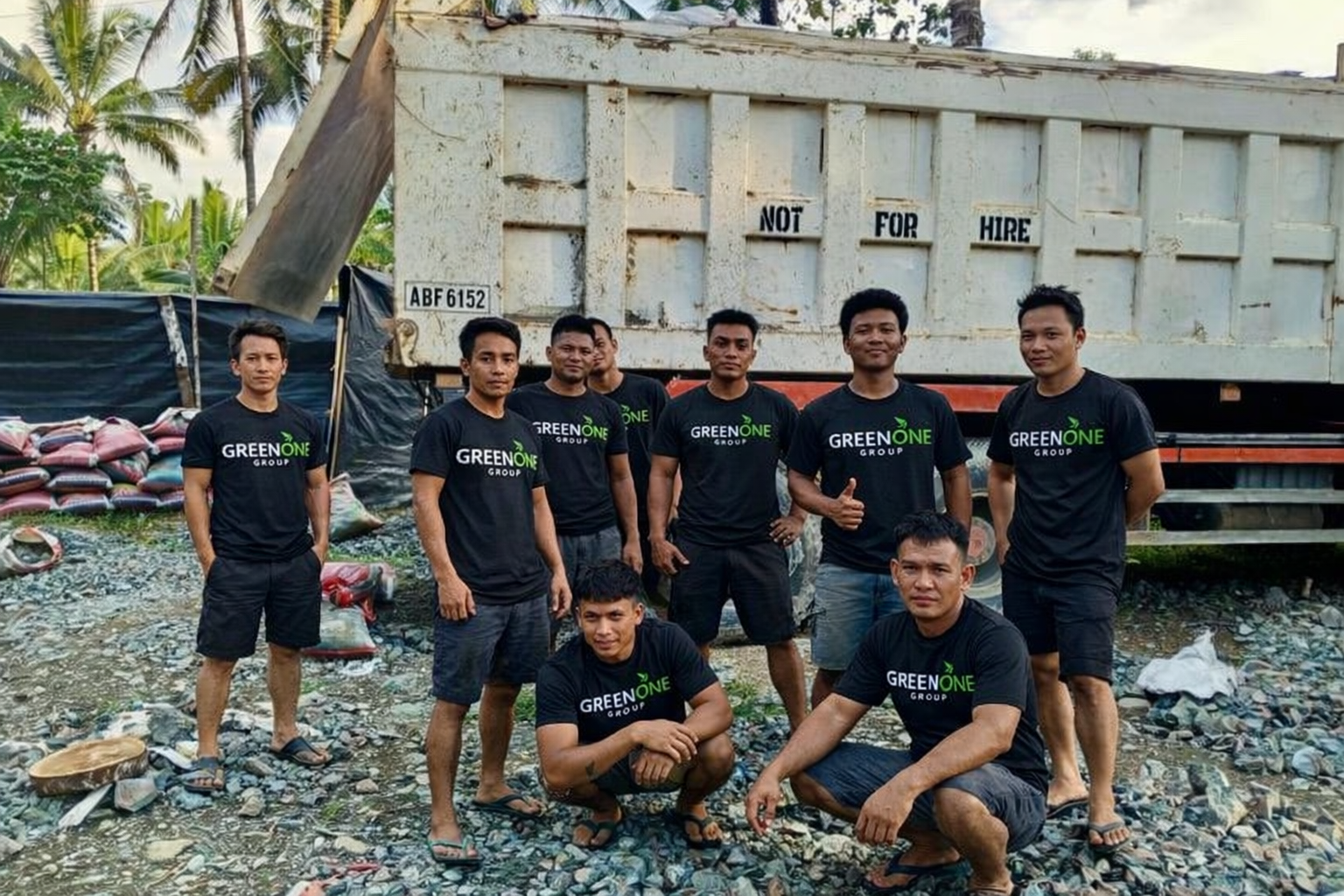 Group of nine men wearing black t-shirts with 'Green One Group' logo, standing and squatting near a large truck on a rocky beach with palm trees in the background.