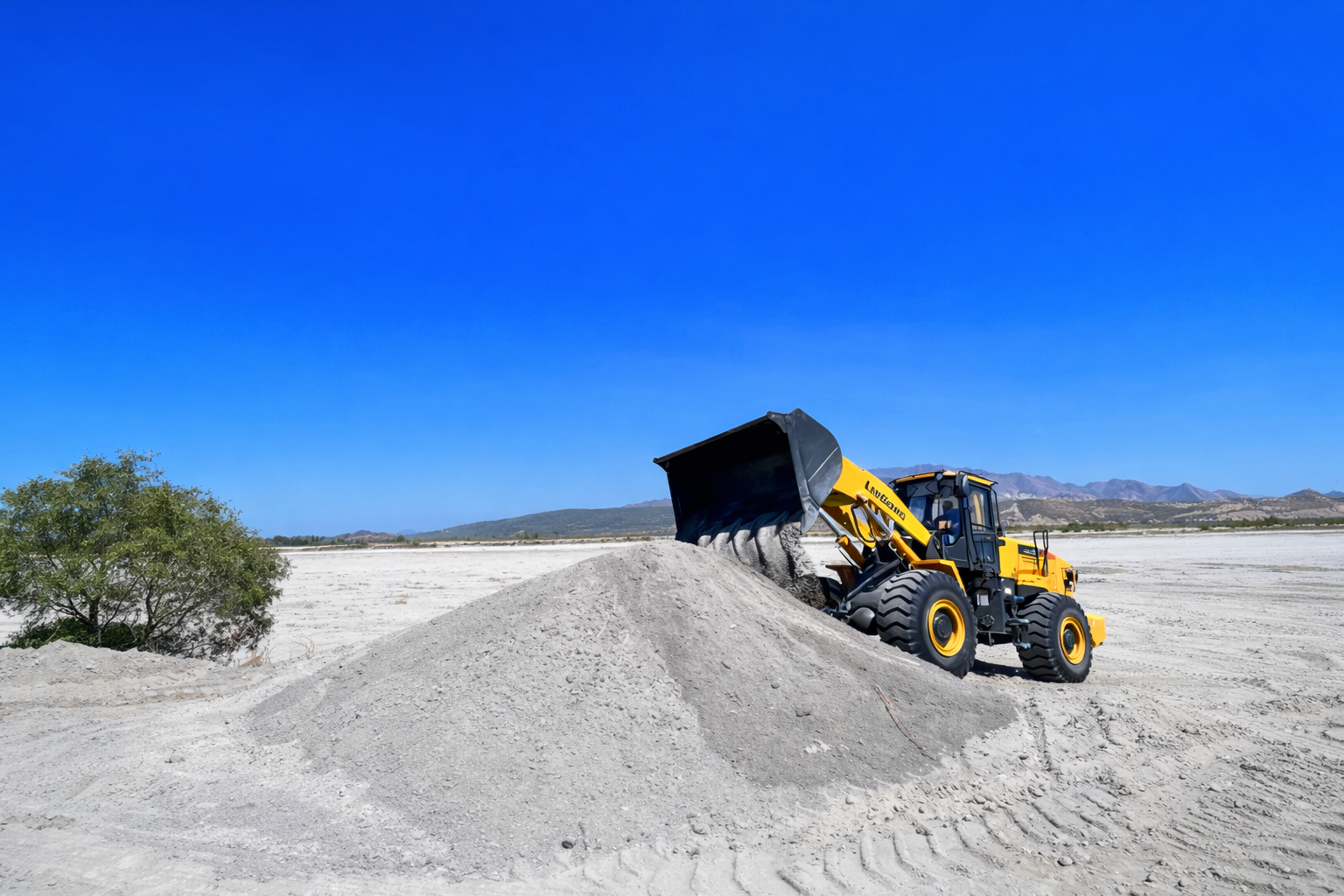 A yellow front loader moving dirt in a barren landscape with mountains in the distance and a clear blue sky.