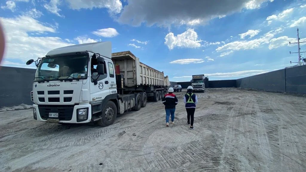 Two construction workers wearing hard hats walking on a dirt construction site, with several large trucks in the background under a blue sky with scattered clouds.